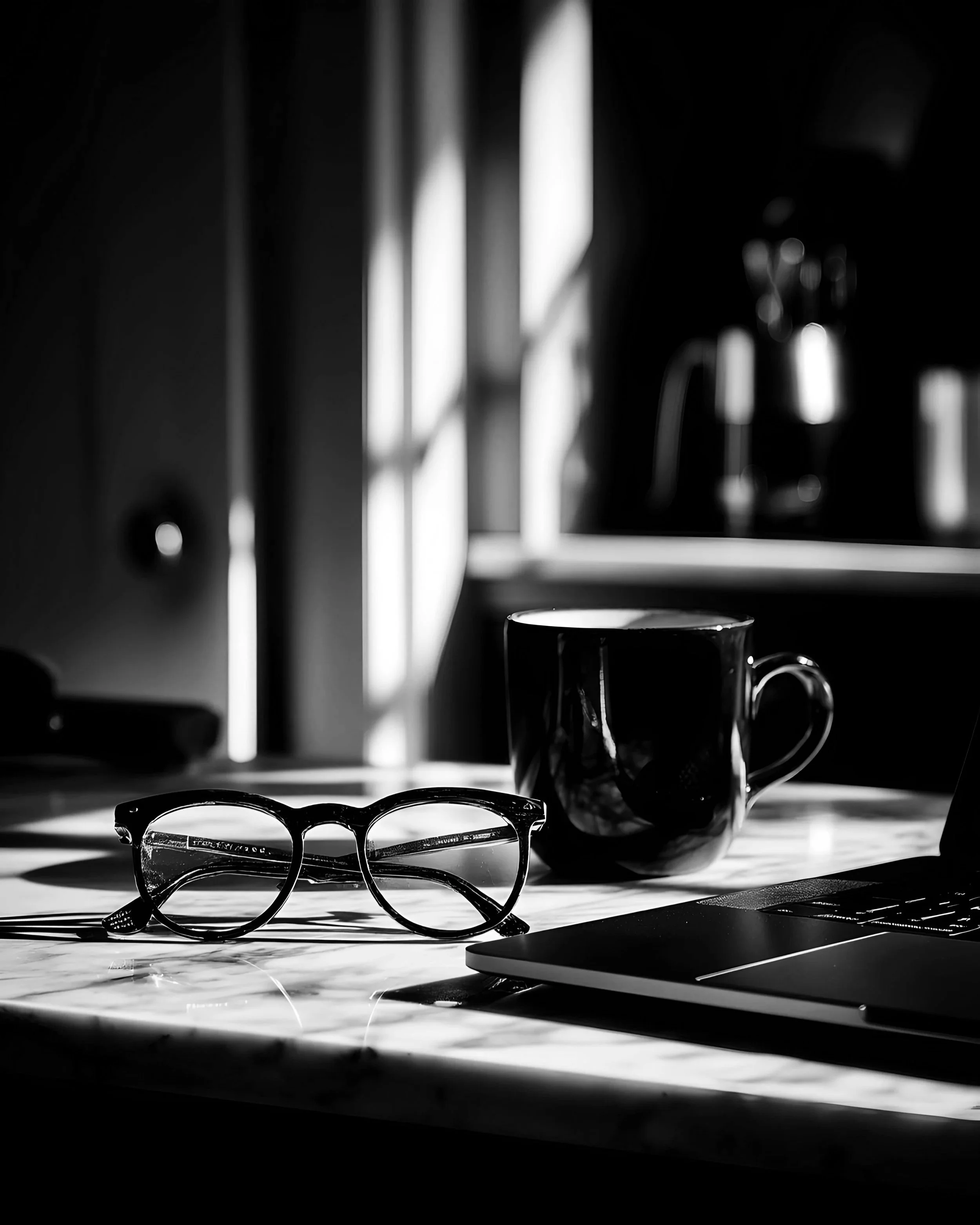 Black and white photo of a desk with eyeglasses, a coffee mug, and a closed laptop, with sunlight and shadow creating a pattern on the surface.