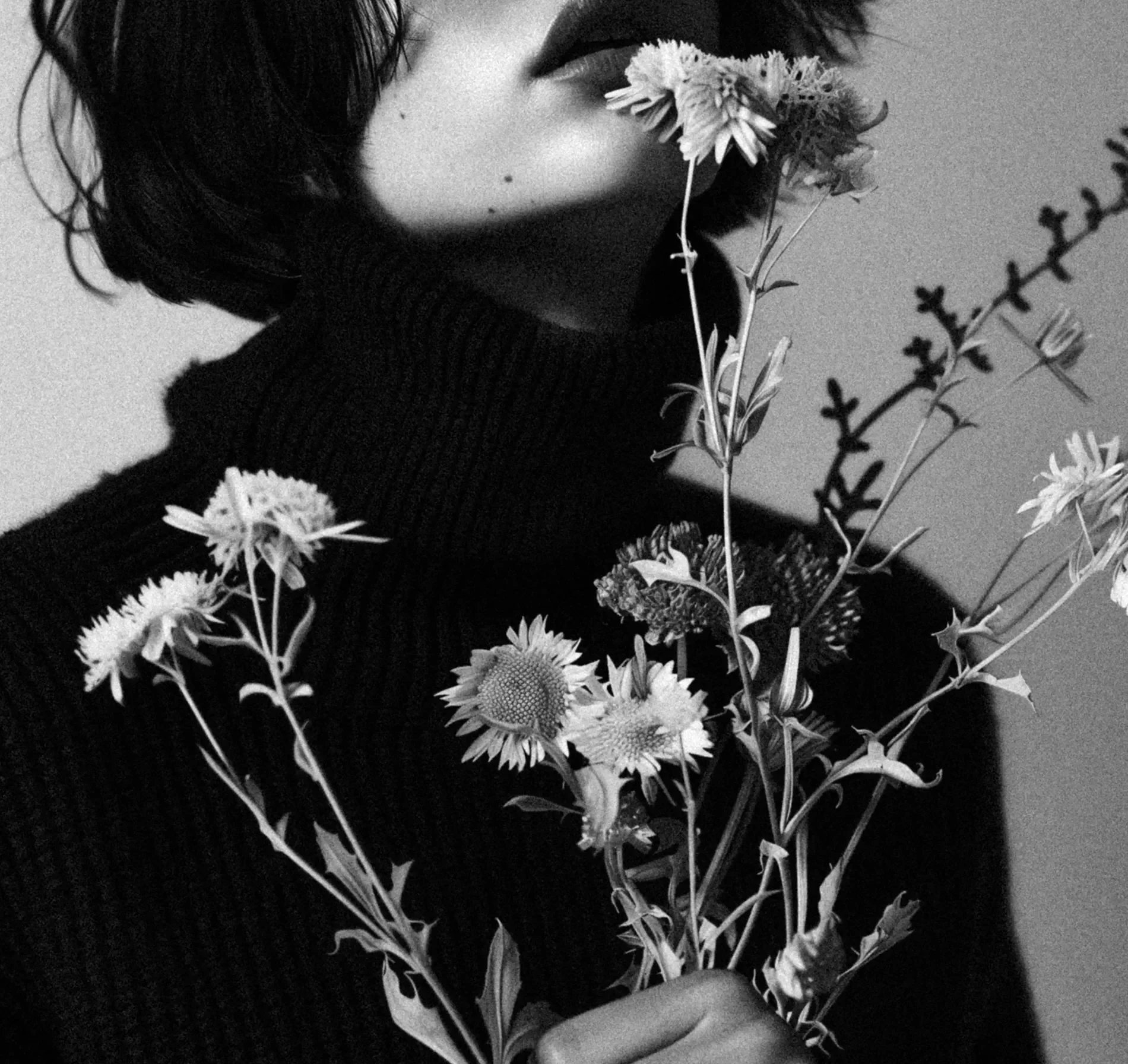 Black-and-white photo of a person with short hair holding a bouquet of flowers close to their face, with their eyes closed and lips slightly parted.