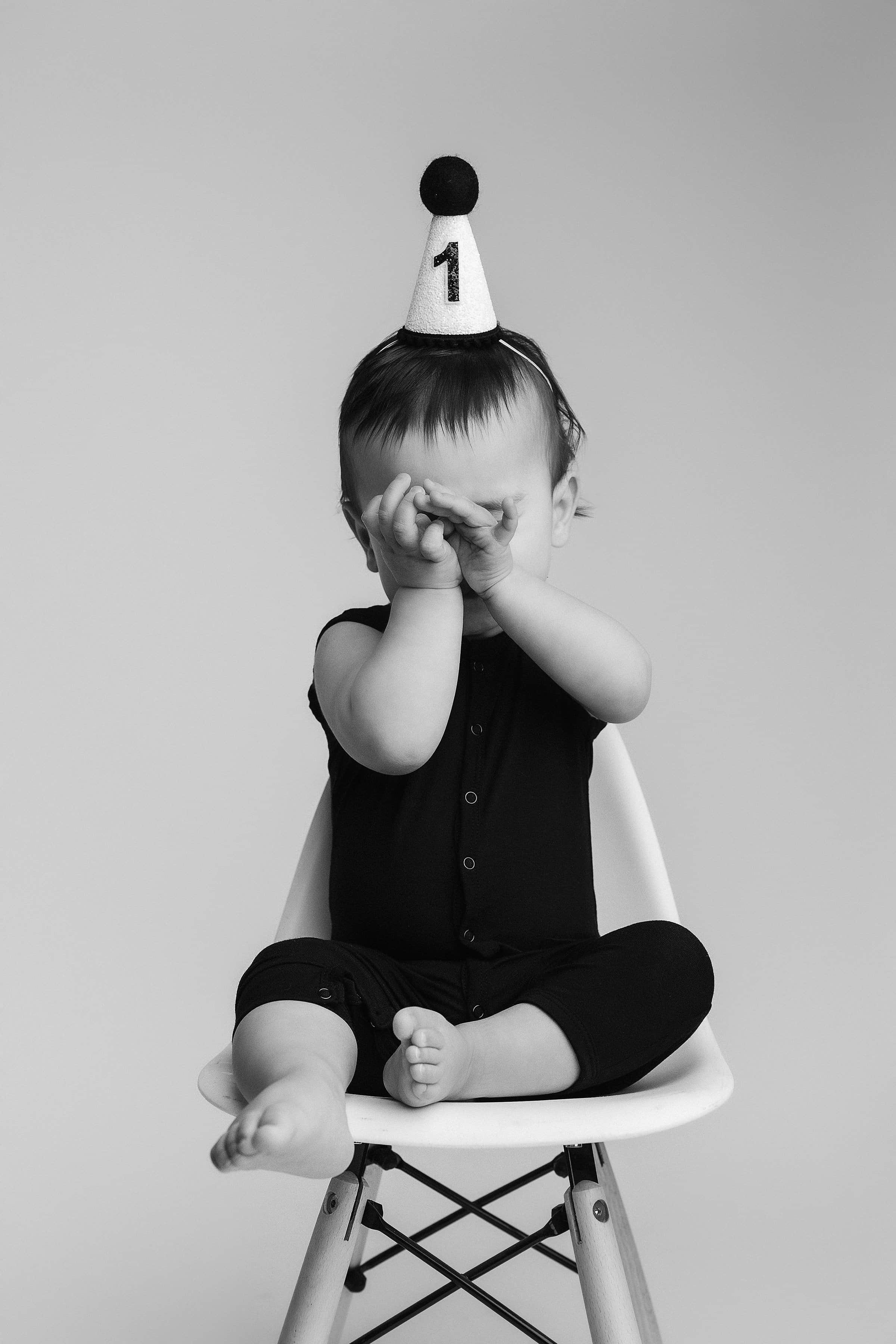 Child sitting on a chair wearing a party hat with the number 1, covering eyes with hand, black and white photo.