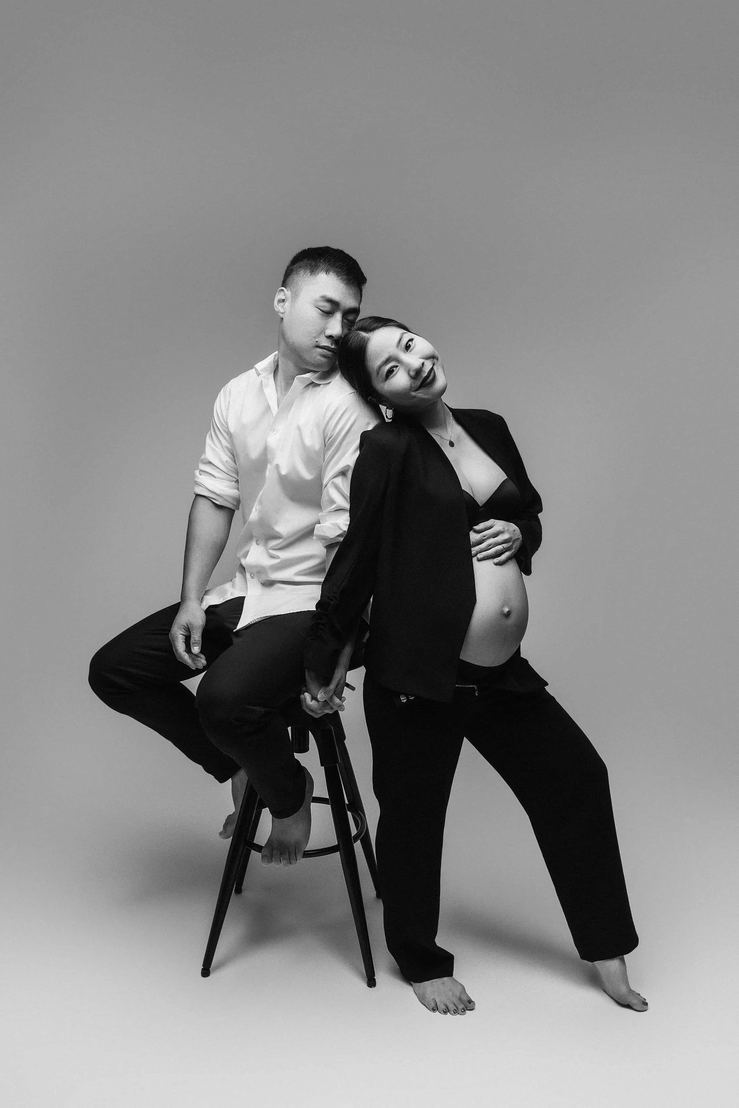 A black and white photo of a pregnant woman with dark hair and a big smile, wearing a black top and pants, standing with her hand on her stomach. She is posing next to a man with short hair and closed eyes, sitting on a stool, wearing a white shirt a