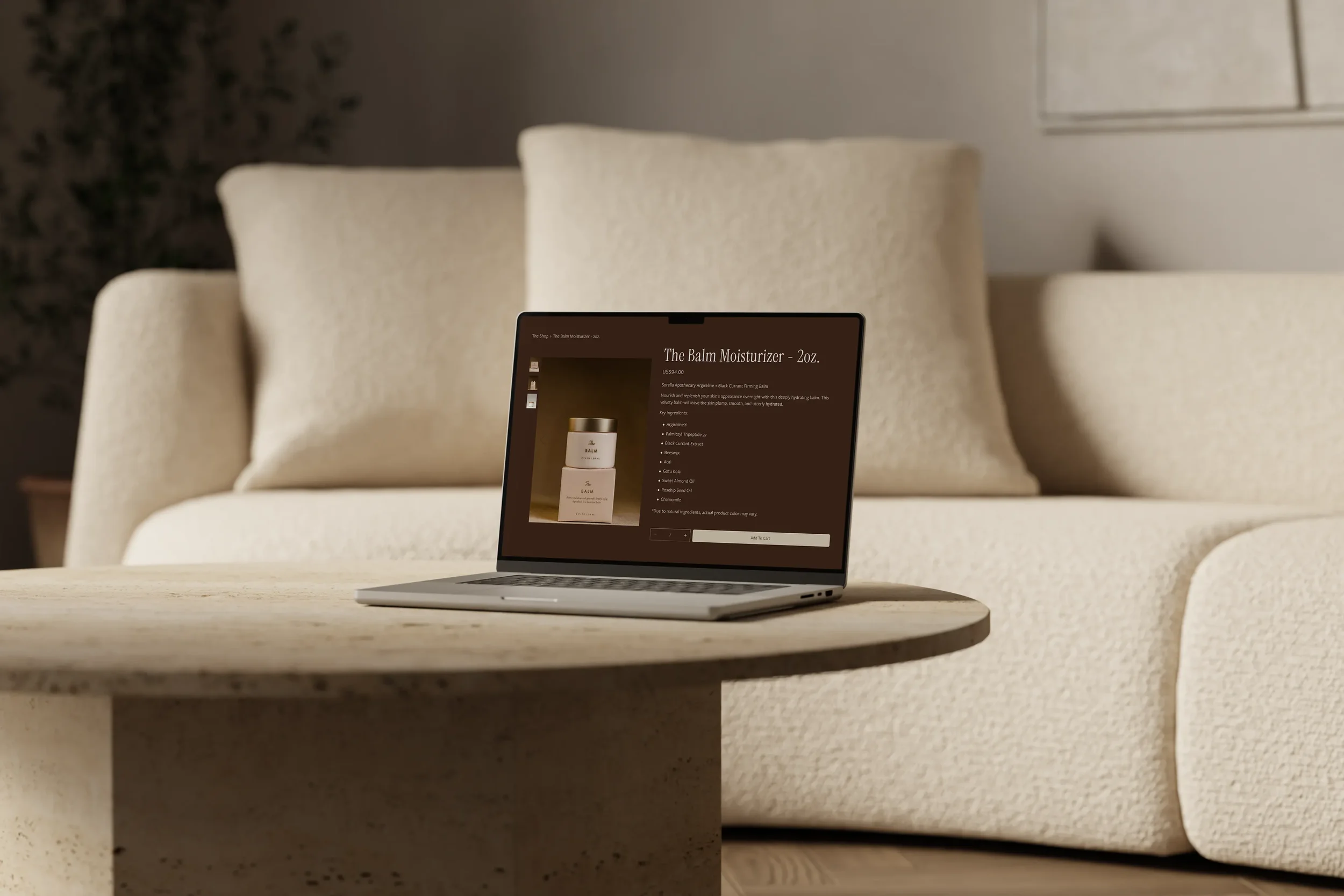 A laptop on a white table displaying an online product page for The Balm Moisturizer, with large beige cushions and a wall in the background.
