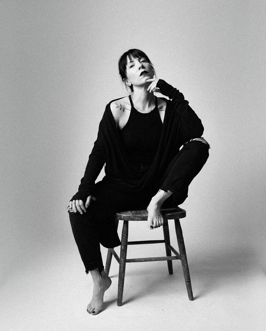 A woman with short dark hair and tattoos sitting on a wooden stool, wearing black clothing, barefoot, with a contemplative expression.