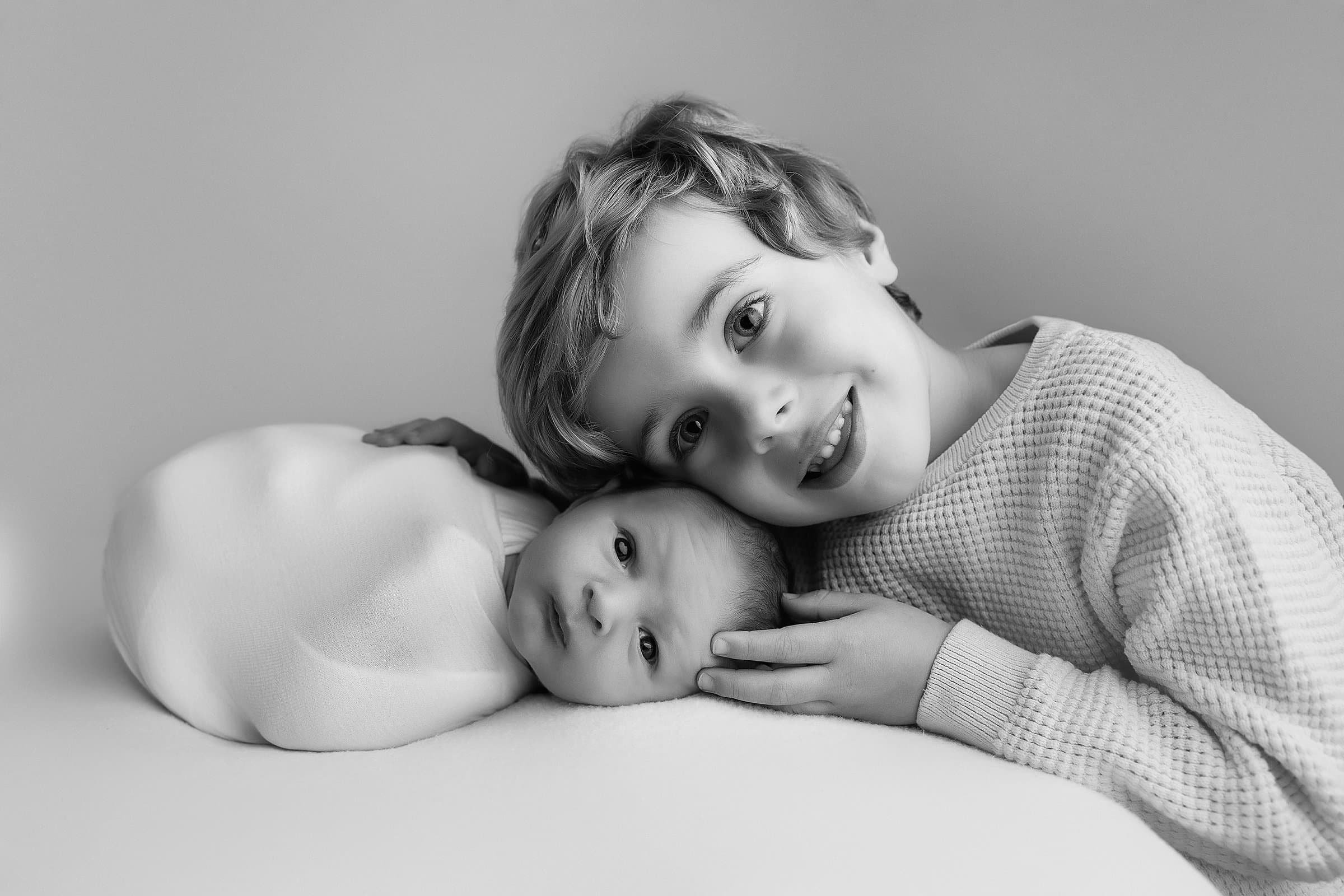 Black and white photograph of three children lying on a bed, with a light-colored wall in the background. The oldest child, a boy with wavy hair, is smiling and resting his head on the bed while embracing two younger children, one with closely croppe