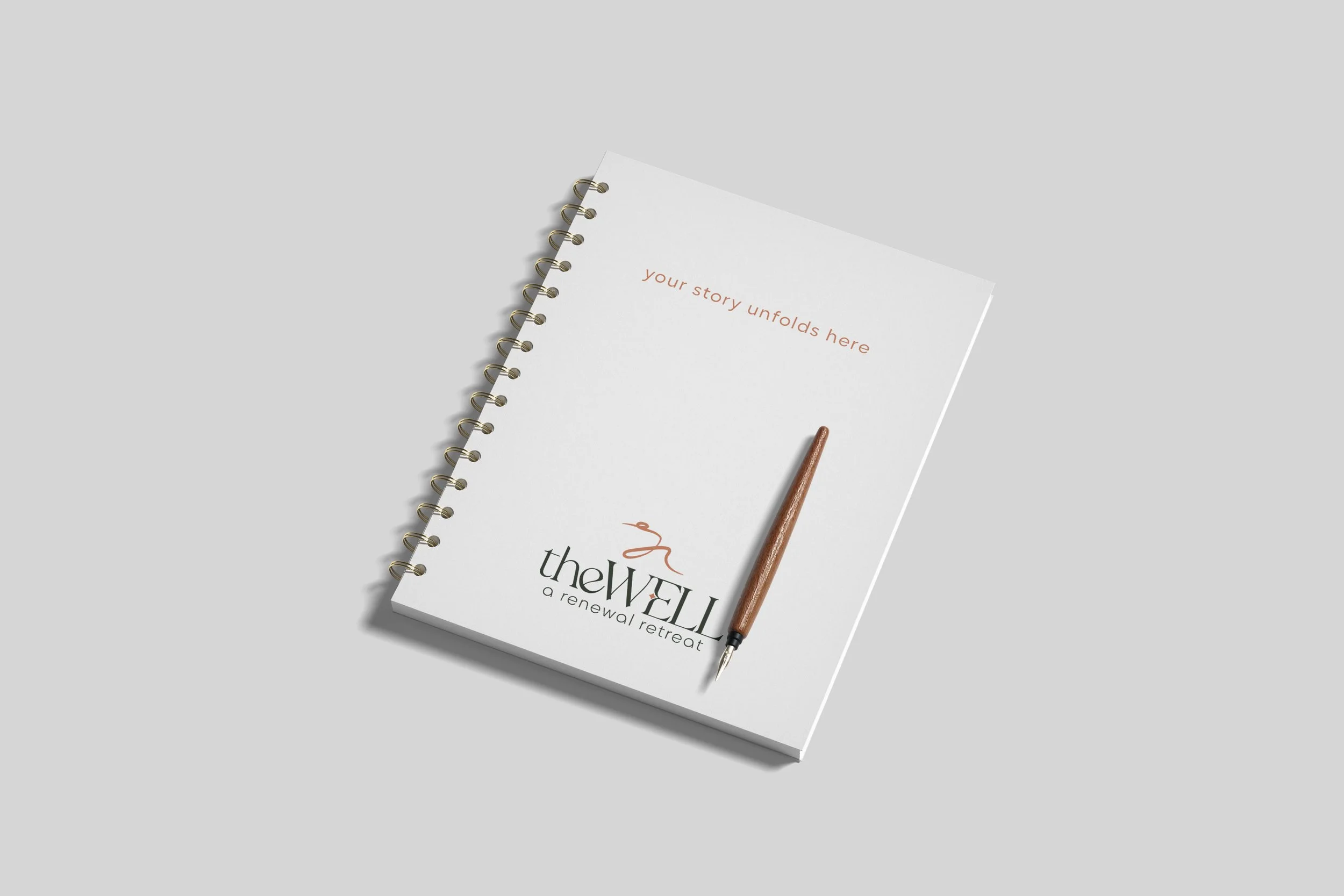 Spiral-bound notebook with the words 'your story unfolds here' and logo for 'the WELI a renewal retreat,' accompanied by a pen on a light gray surface.