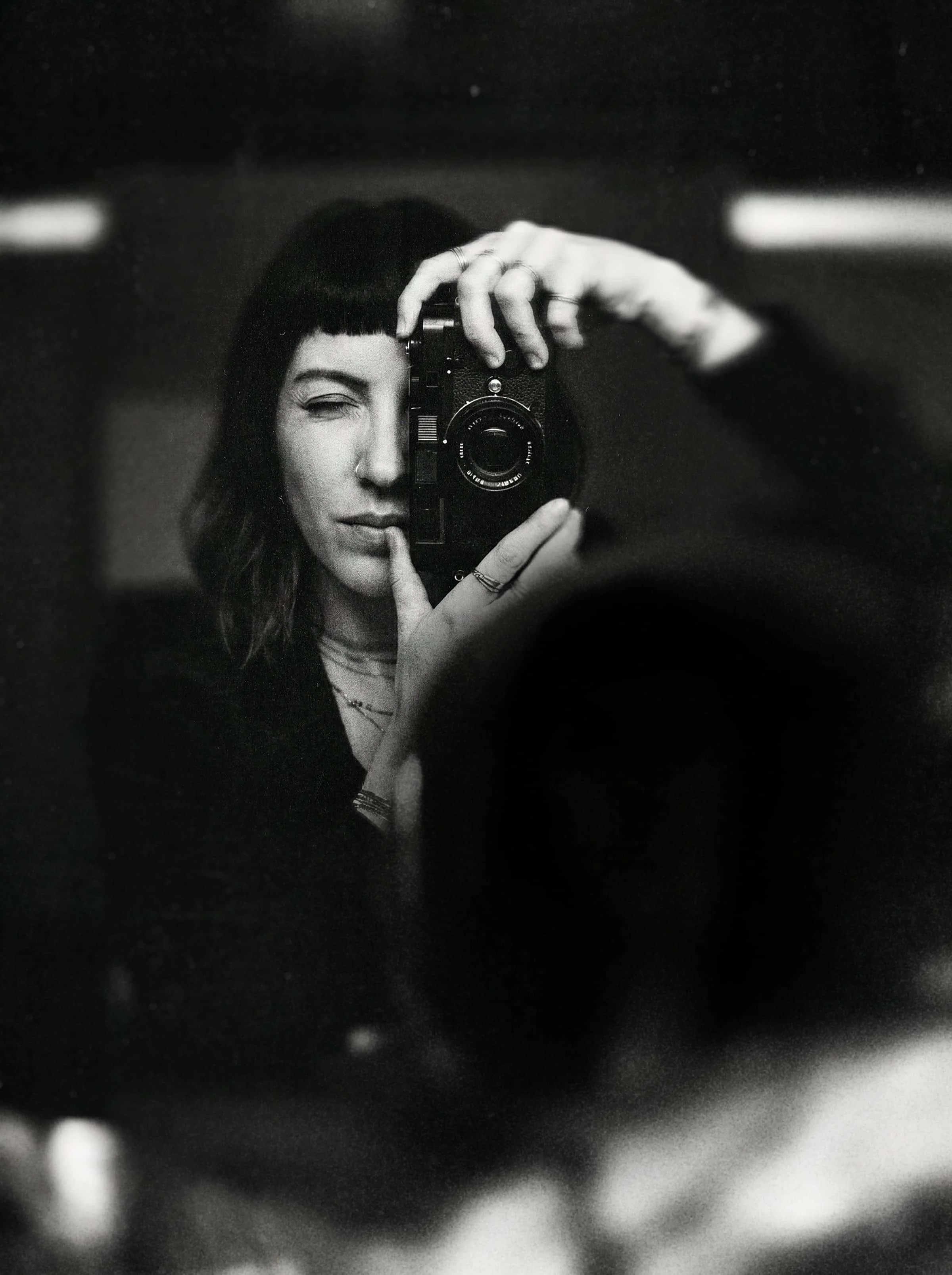 A person with dark hair and a nose piercing taking a selfie in a mirror with a camera. The photo is in black and white.