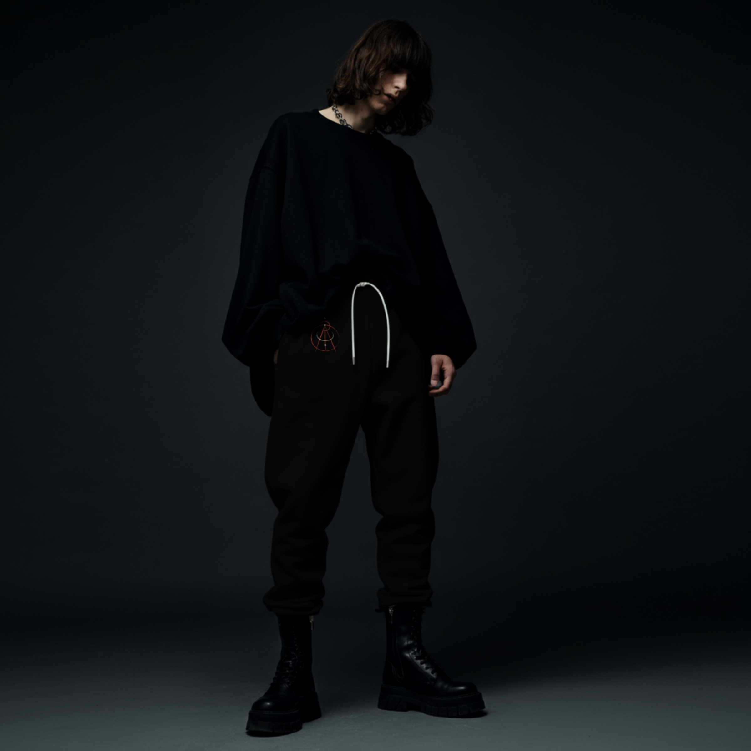 A person standing against a dark background, wearing a loose black sweatshirt, black pants with a white drawstring, and black combat boots with thick soles.