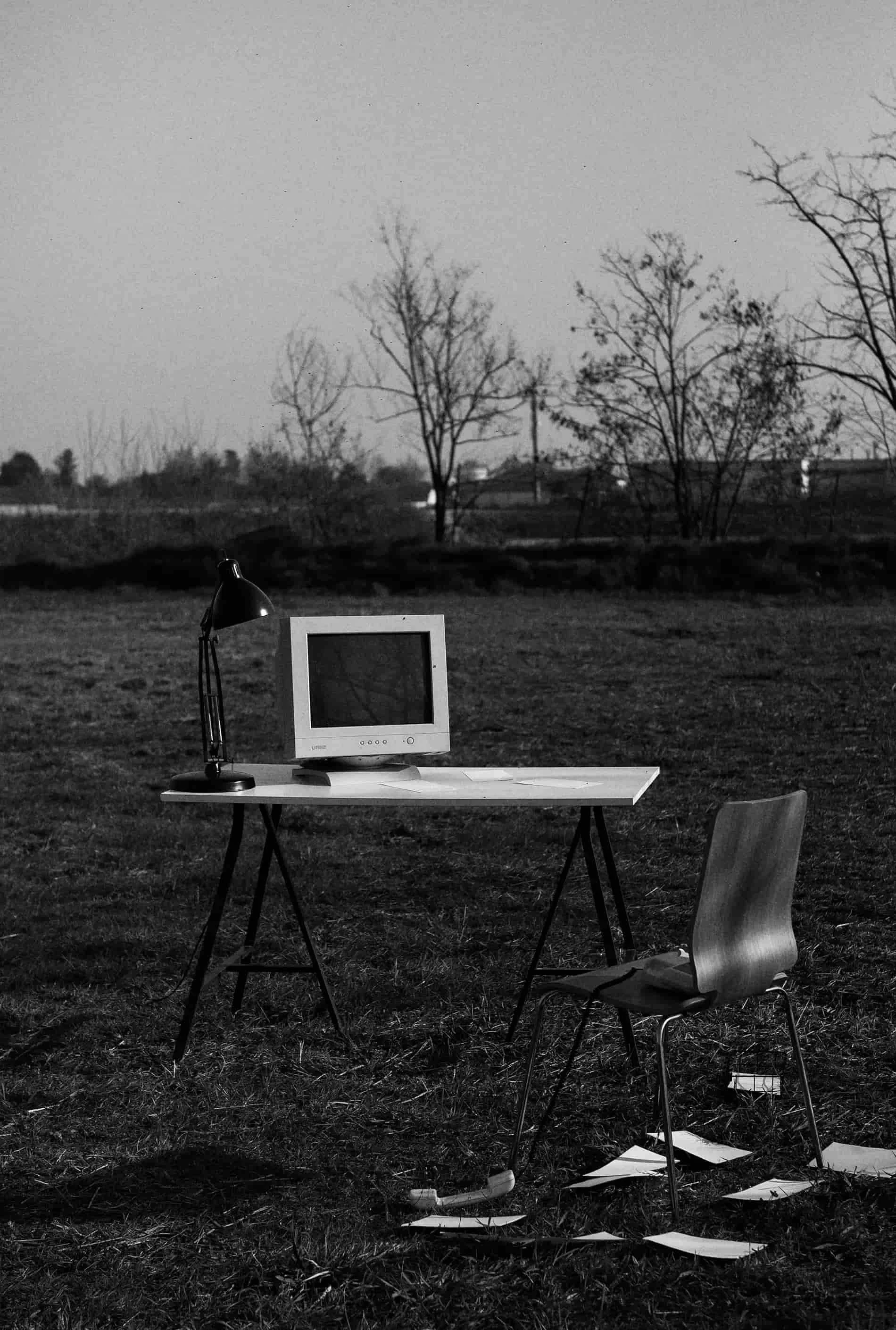 A black and white photograph of an outdoor scene with a desk and chair on a grassy field. The desk has a computer monitor, a desk lamp, and some papers. The chair is positioned near the desk. In the background, there are leafless trees and a cloudy sky.