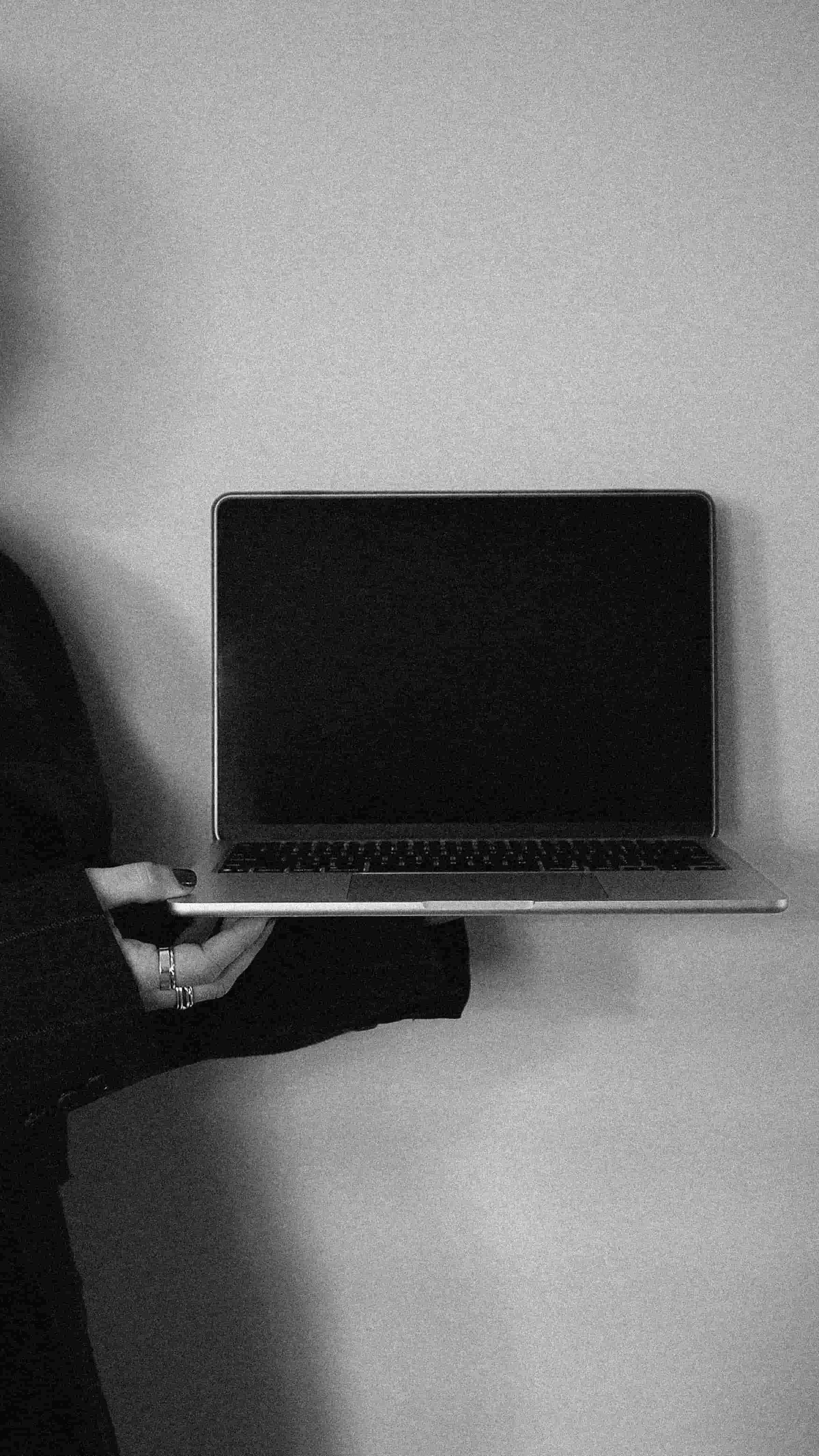 A person holding a laptop with a blank black screen, sitting at a table with a plain background.