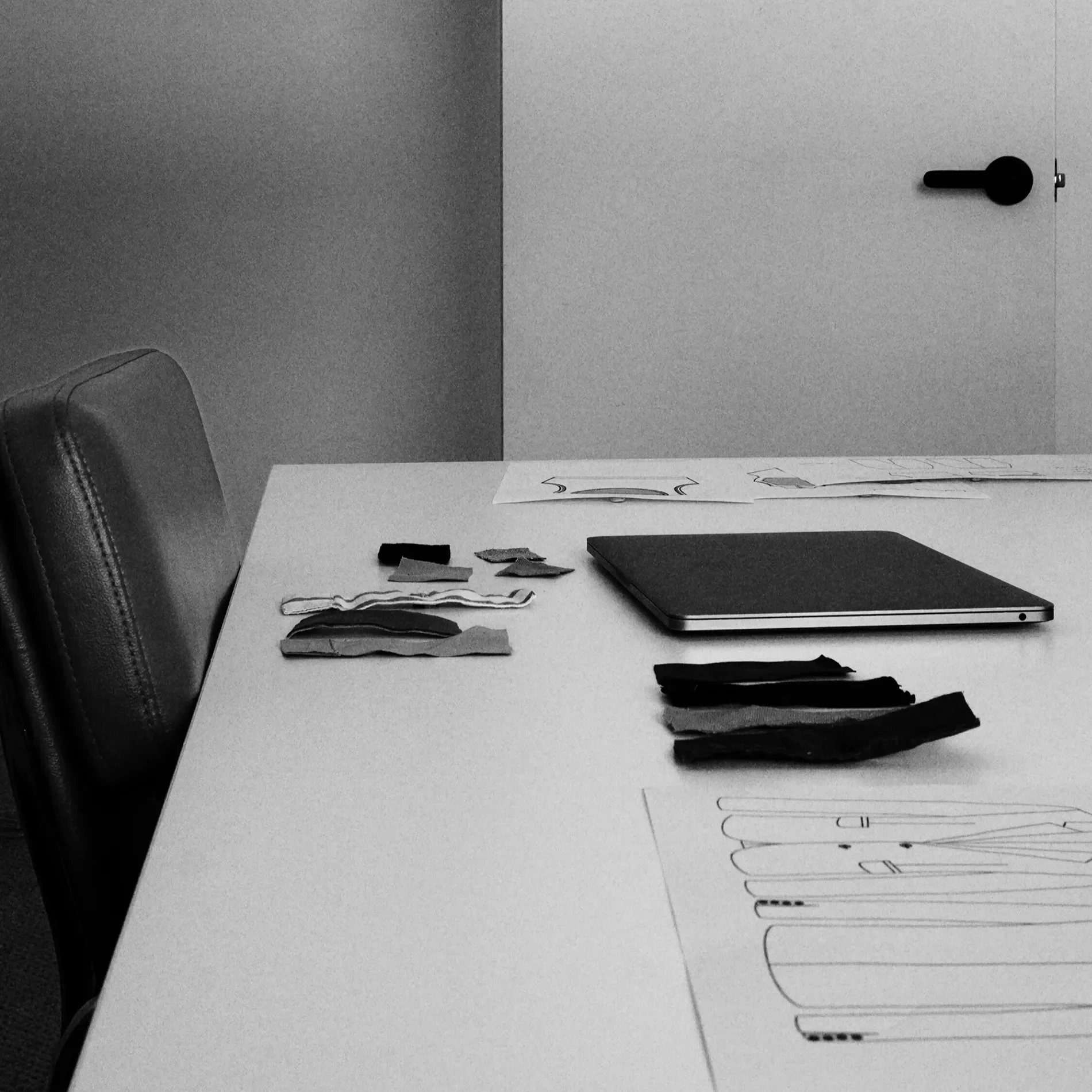 A workspace with a white table, a closed laptop, small pieces of fabric or paper, and drawings or sketches on white sheets. A black chair is partially visible on the left.