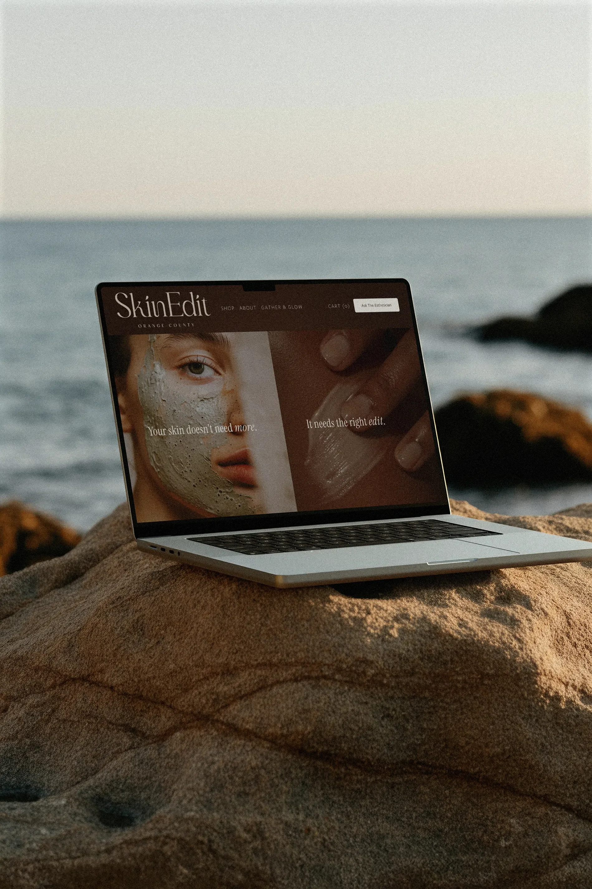 A laptop on a rock near the ocean, displaying a Squarespace skincare website with a woman's face and text about skin care.