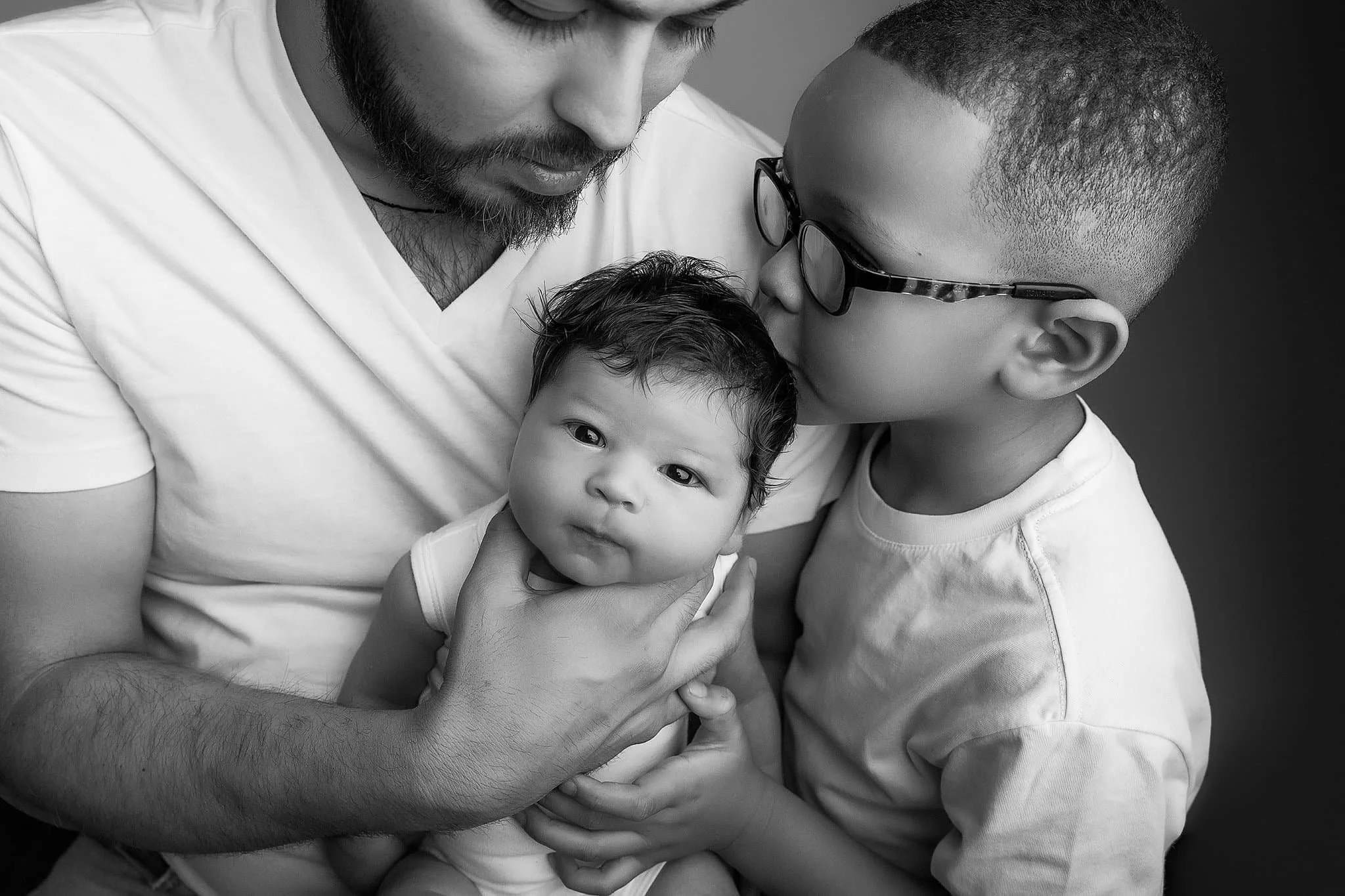 A black-and-white photo of a man with a beard holding and cuddling two young children, one with glasses and short hair kissing the other with curly hair on the forehead.