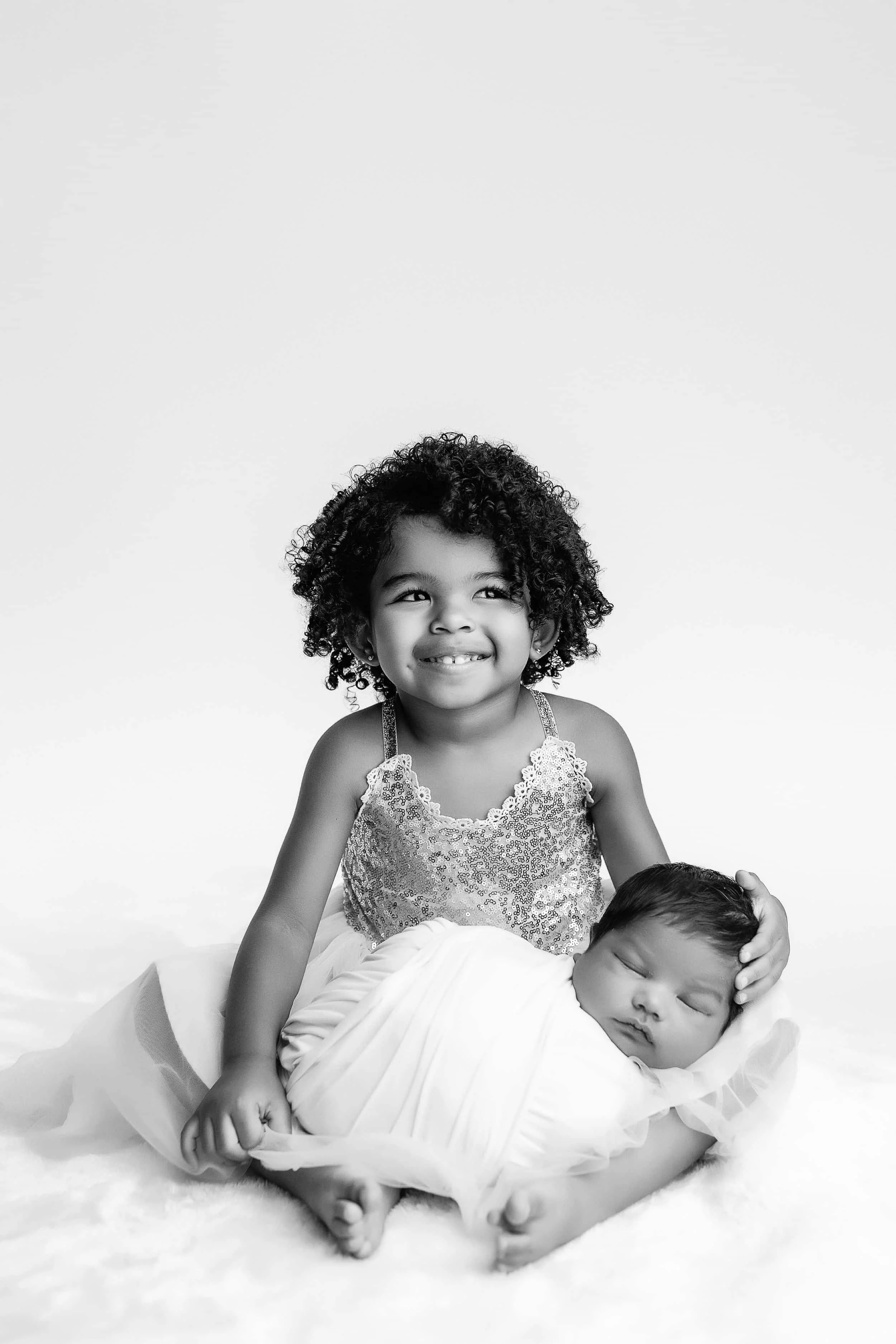 A young girl with curly hair and a bright smile, wearing a sequin dress, is sitting and looking to the side. She is holding a sleeping baby wrapped in a white blanket, laying on her lap, on a soft white surface.