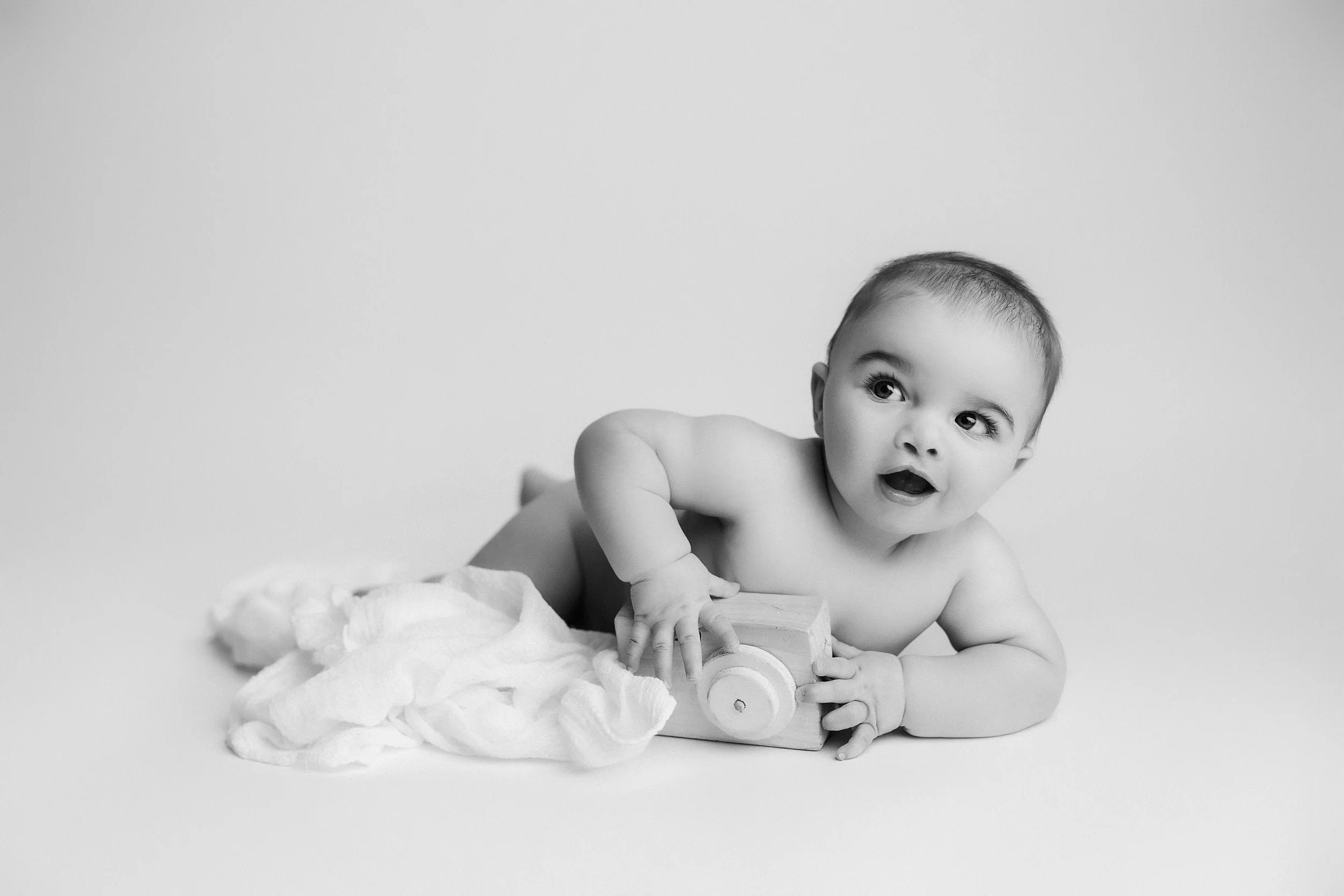 A black-and-white photo of a baby lying on a soft blanket, holding a toy, with a surprised expression, against a plain background.