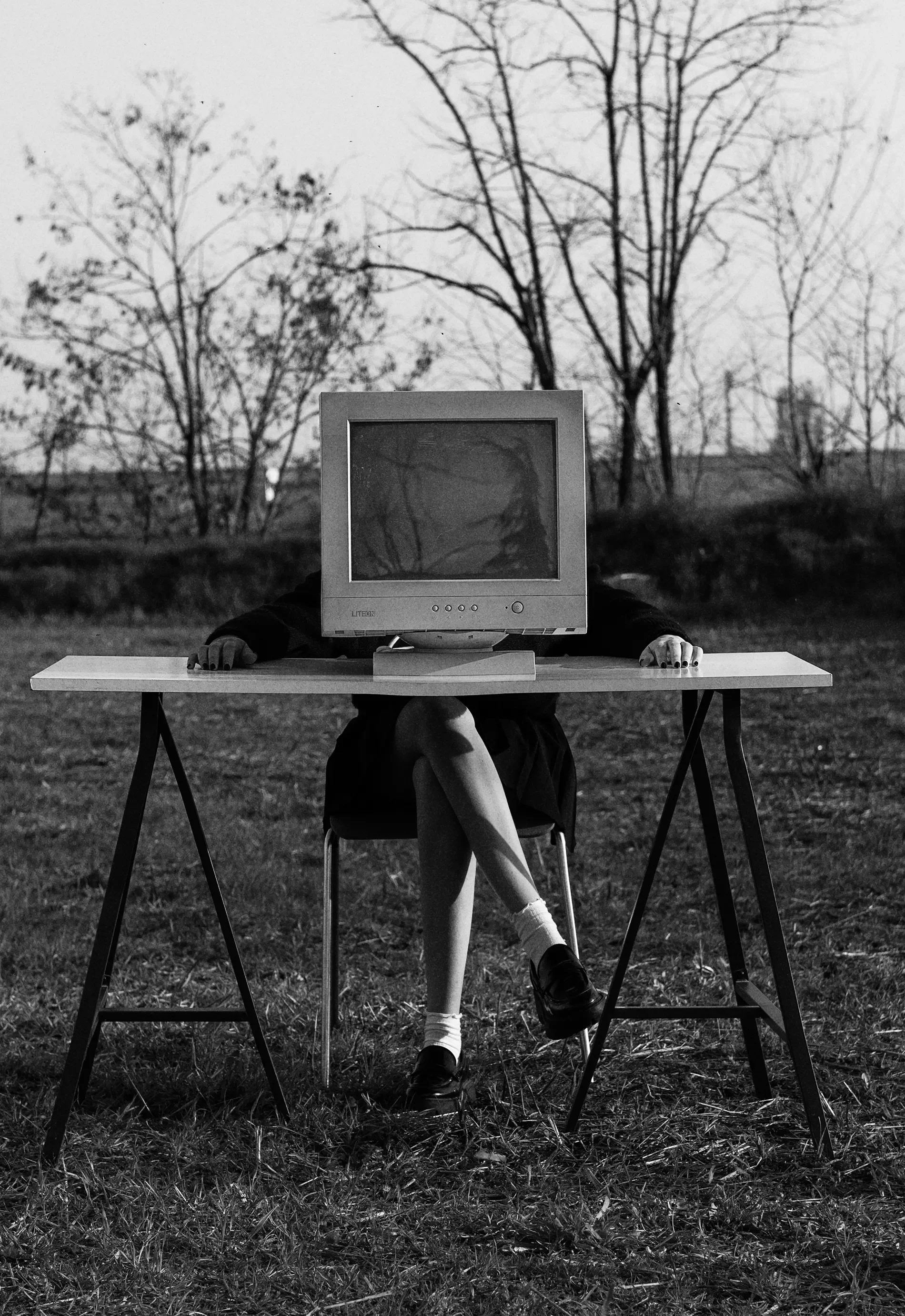 A person with a computer monitor for a head sitting at a table outdoors. The person is wearing a long-sleeved shirt, a skirt, and black shoes. Their hands are resting on the table, and their legs are crossed. The background features leafless trees and a cloudy sky.