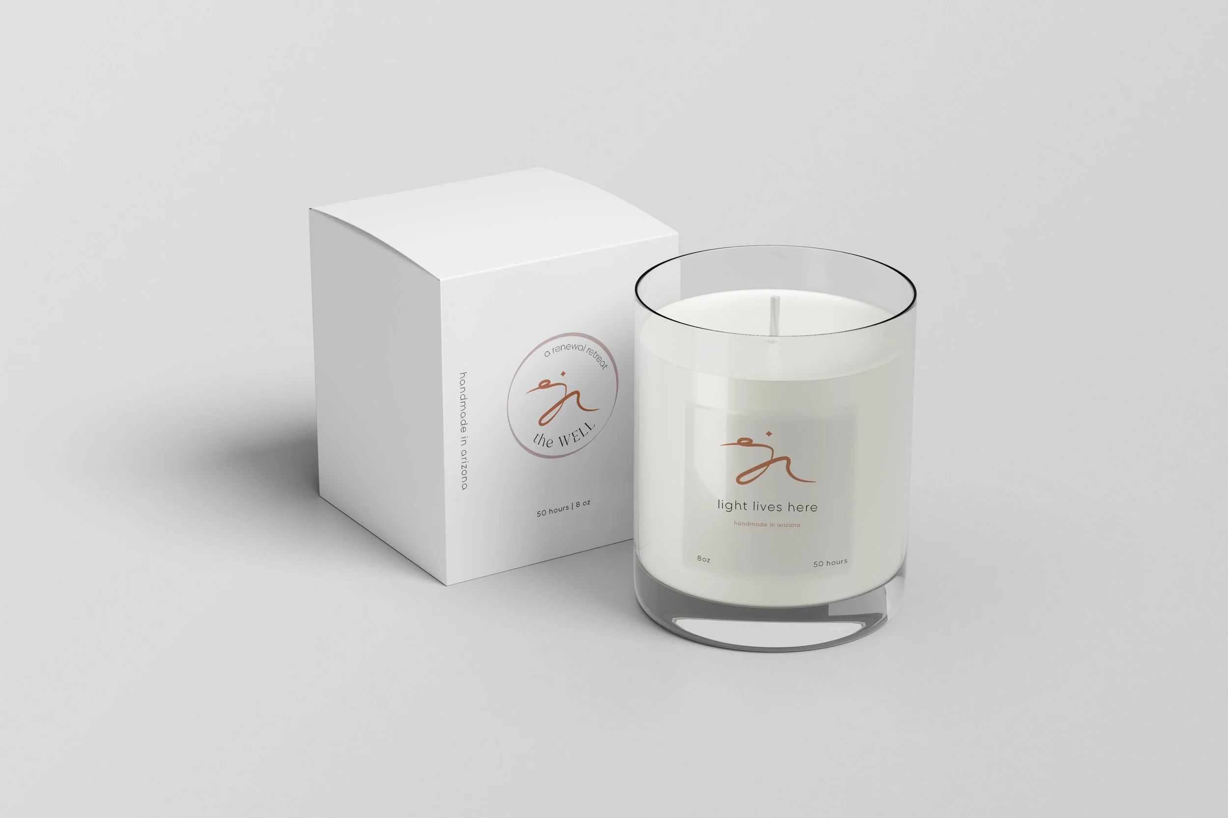 A white candle in a clear glass holder next to a white box with minimalist design, branding 'the WELL', and the text 'light lives here' and 'handmade in arizona'.