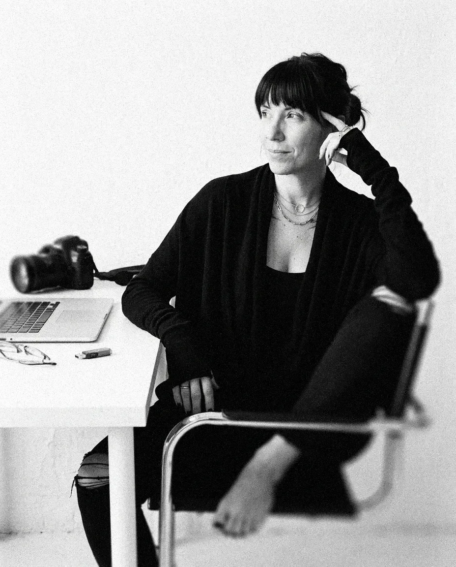 Black-and-white photo of a woman sitting at a desk, looking thoughtfully to the side. There is a camera and a laptop on the desk.