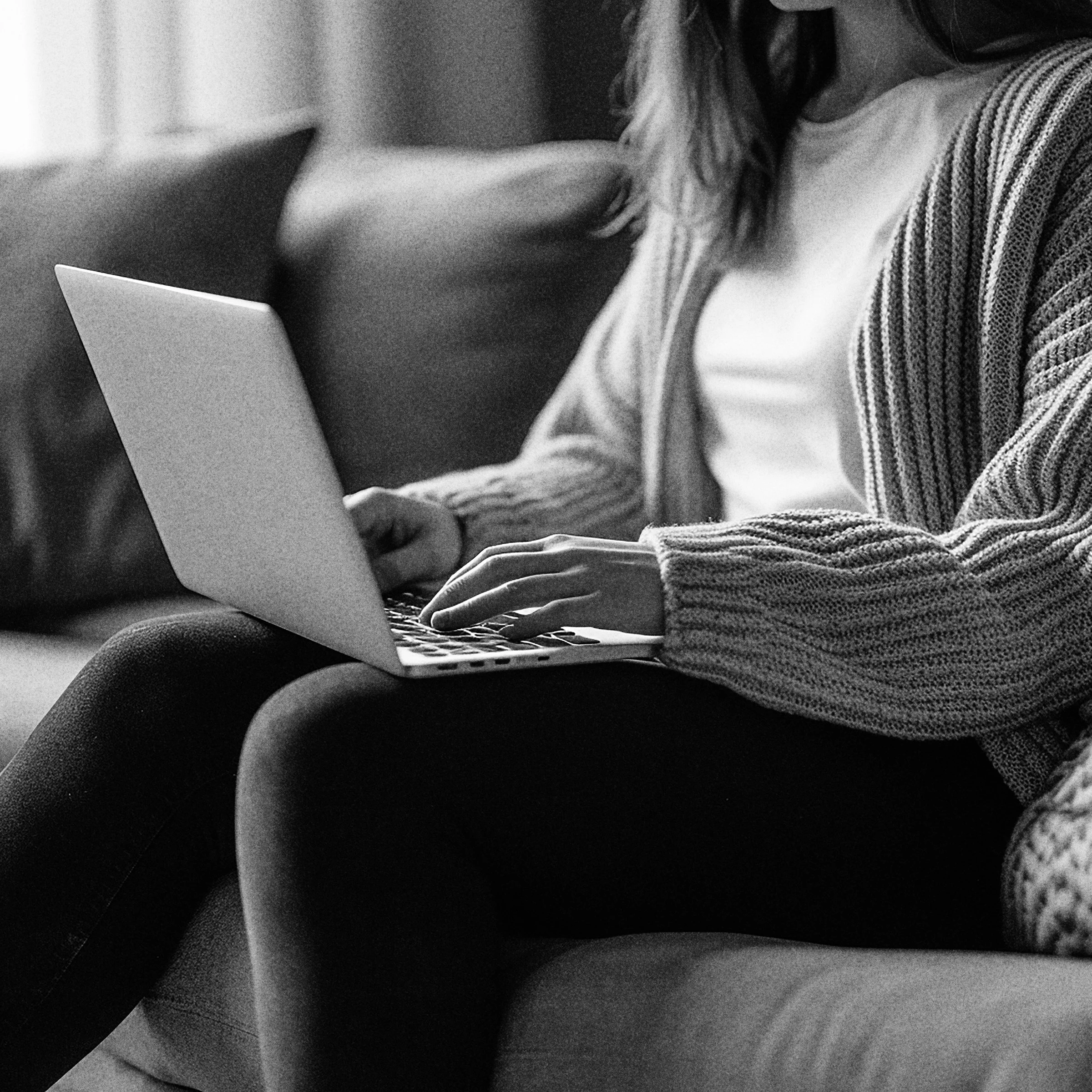 Person sitting on a sofa, using a laptop, wearing a striped cardigan and dark pants, in a black and white photo.