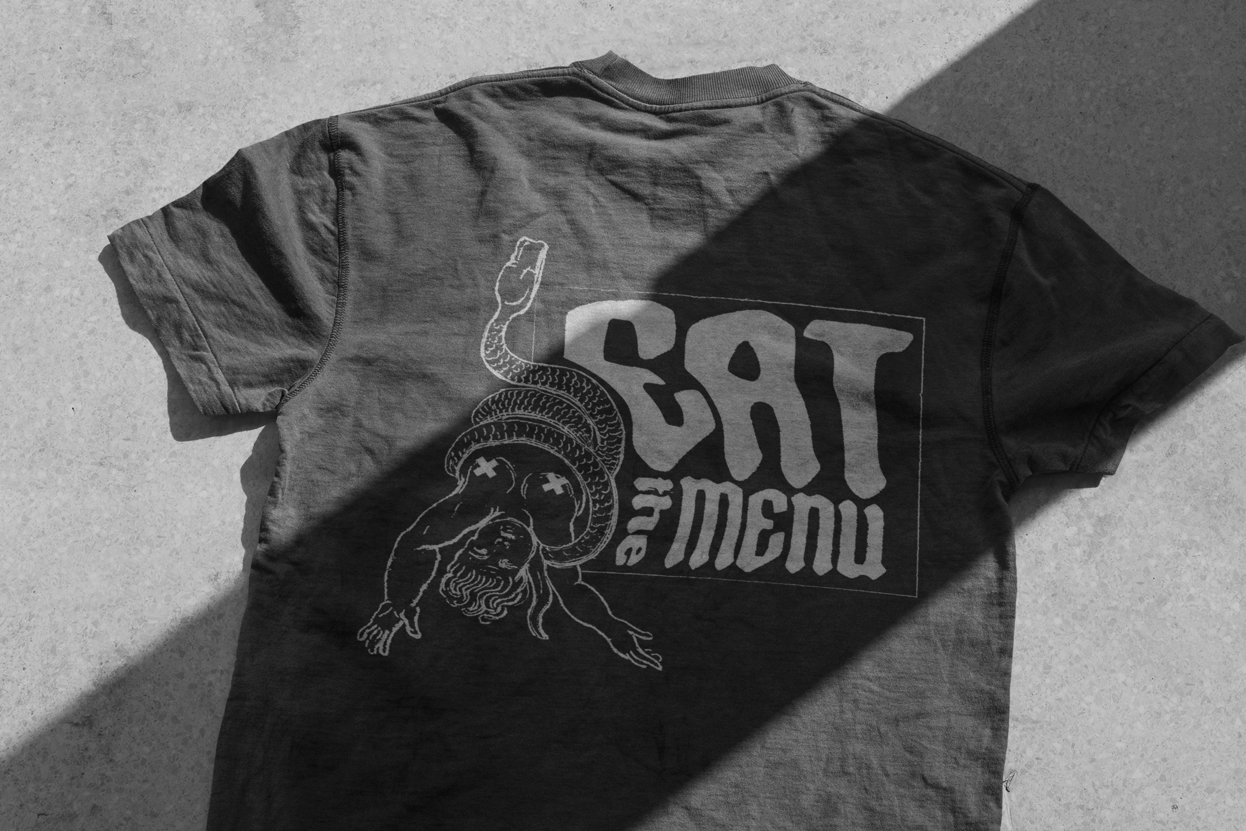 A gray t-shirt with a graphic design featuring a snake and a muscular figure, laid flat on a concrete surface with a shadow falling across the top right corner.