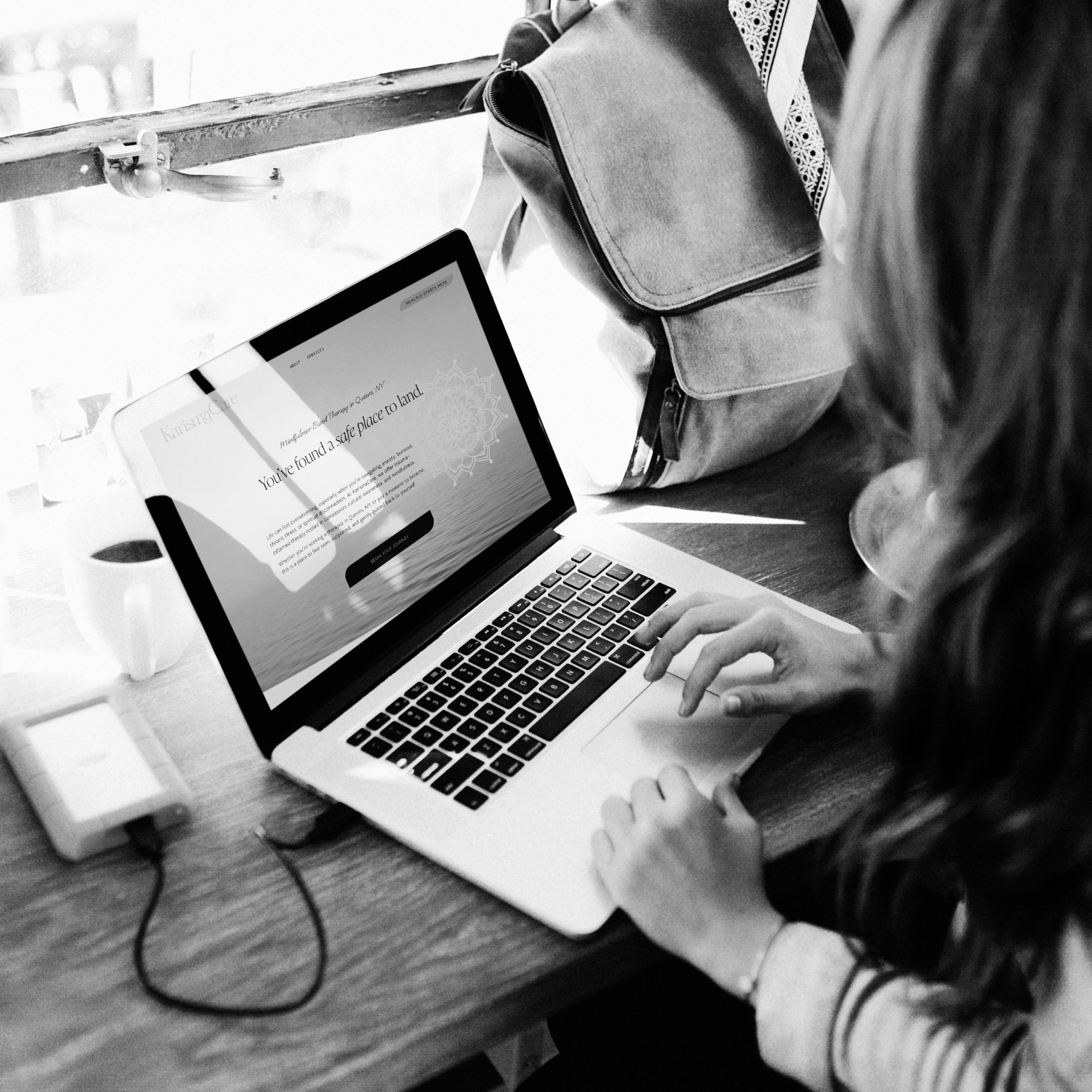 Person using a laptop at a table in a cafe. The laptop screen displays a message: 'You've found a safe place to land.' A coffee cup, portable charger, and backpack are on the table.