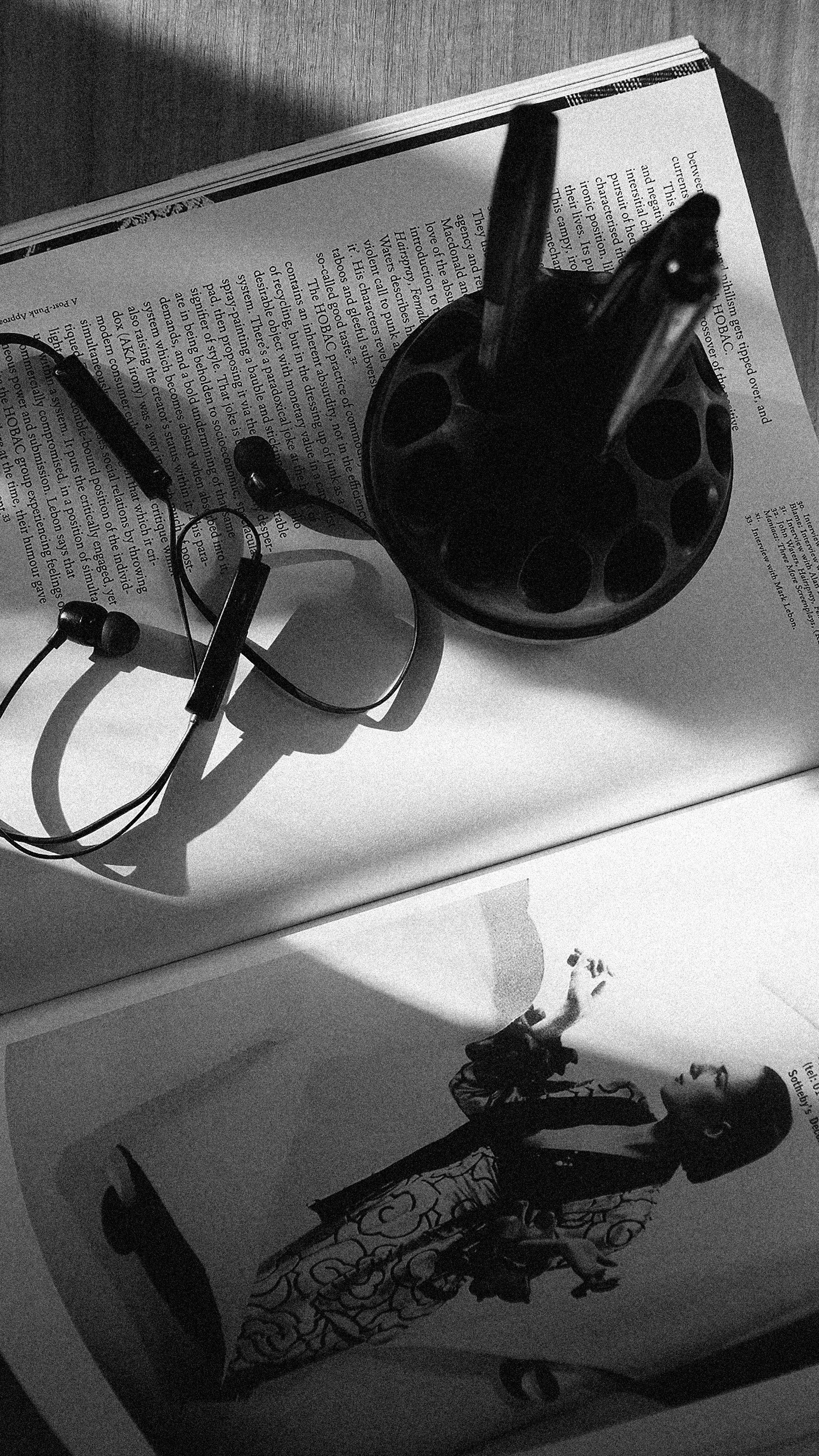 Black and white image of a desk with a book, headphones, a large item resembling a mask or sculpture, and a woman in patterned pants standing nearby, with a small dog at her feet.