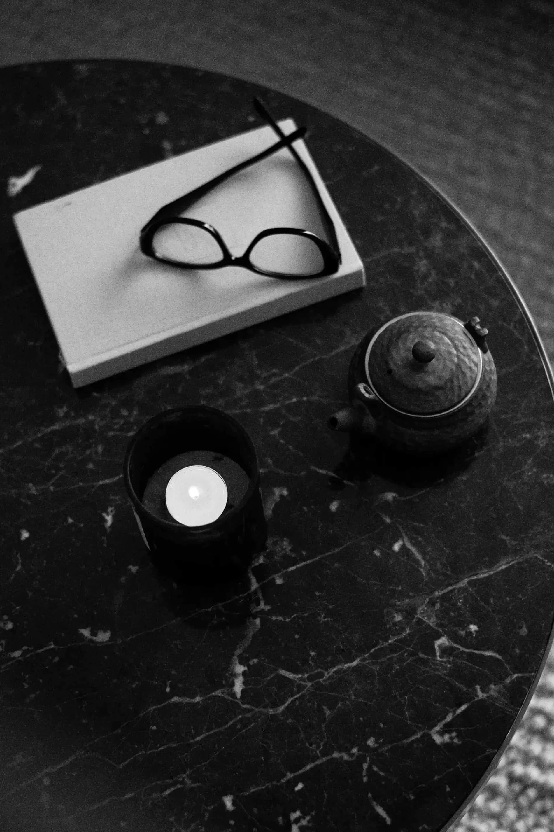 A pair of eyeglasses resting on a white piece of paper on a dark marble surface. Nearby are a small candle holder with a lit candle and a teapot.