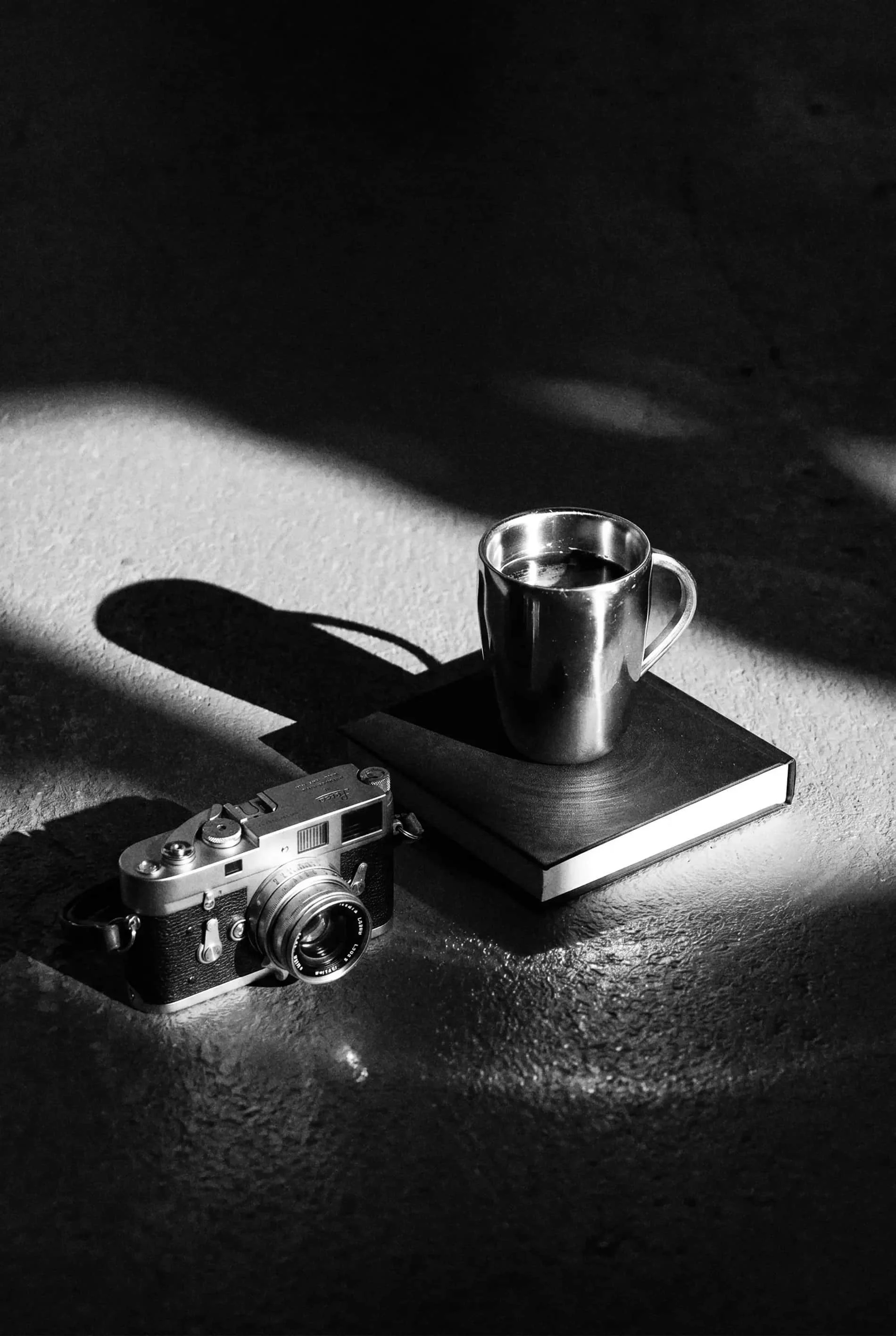 A metal mug filled with coffee, placed on a hardcover book, with an old camera and a matchbox nearby. The scene is illuminated by a bright light creating shadows on the textured surface.