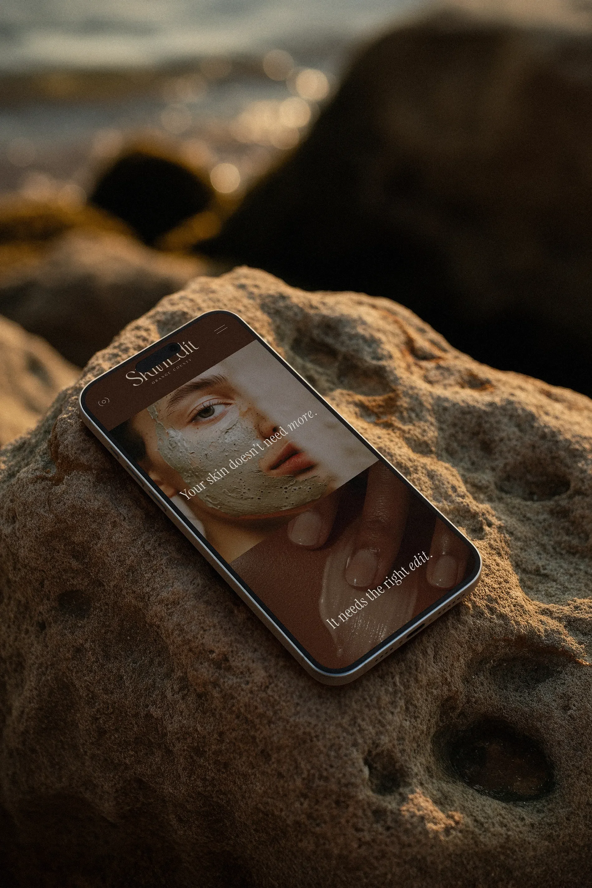A smartphone resting on a rocky surface displays a skincare website with an image of a woman's face with a facial mask and some text overlay.