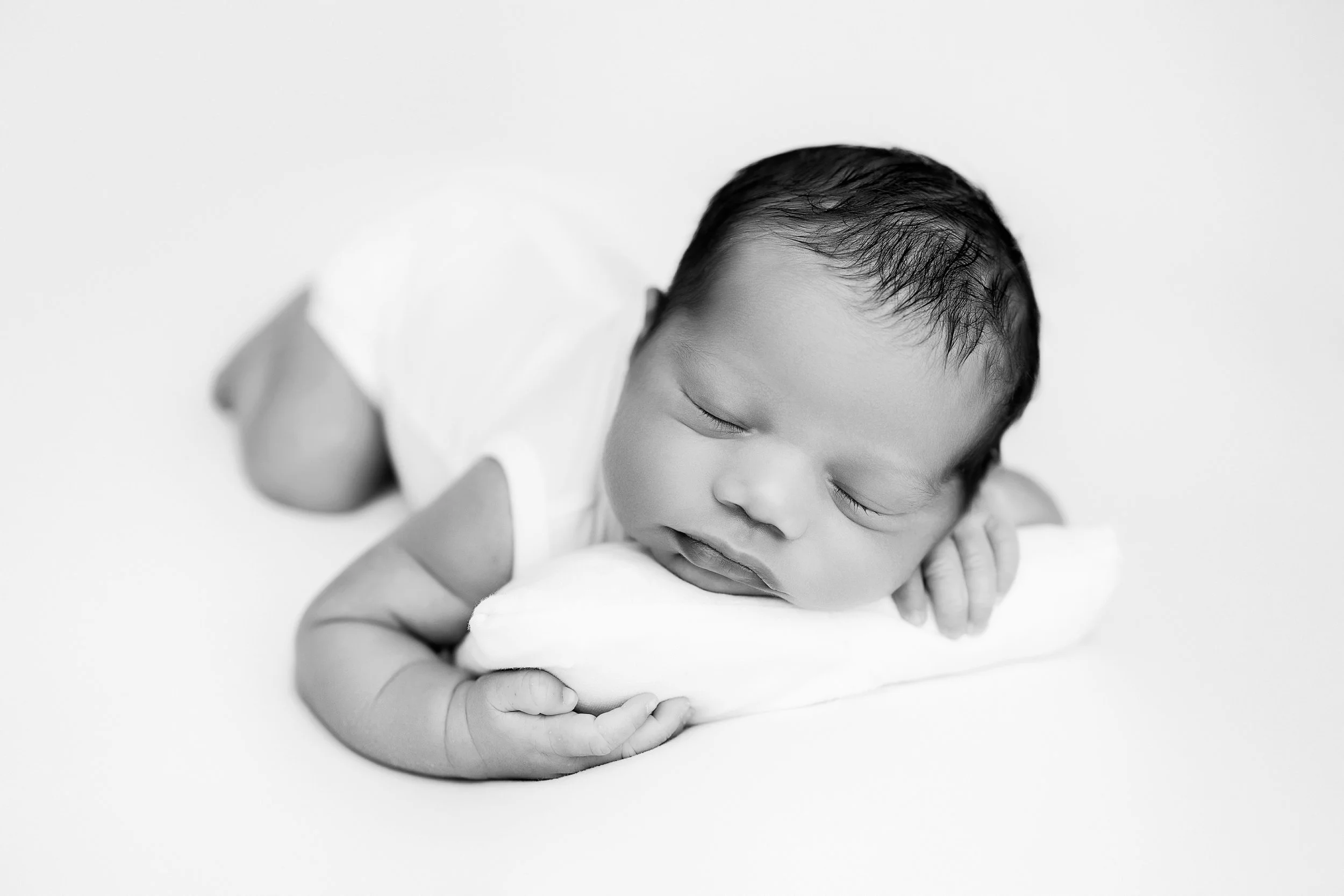 Close-up black and white photograph of a sleeping newborn baby with wet hair, resting its head on its arms, on a plain background.