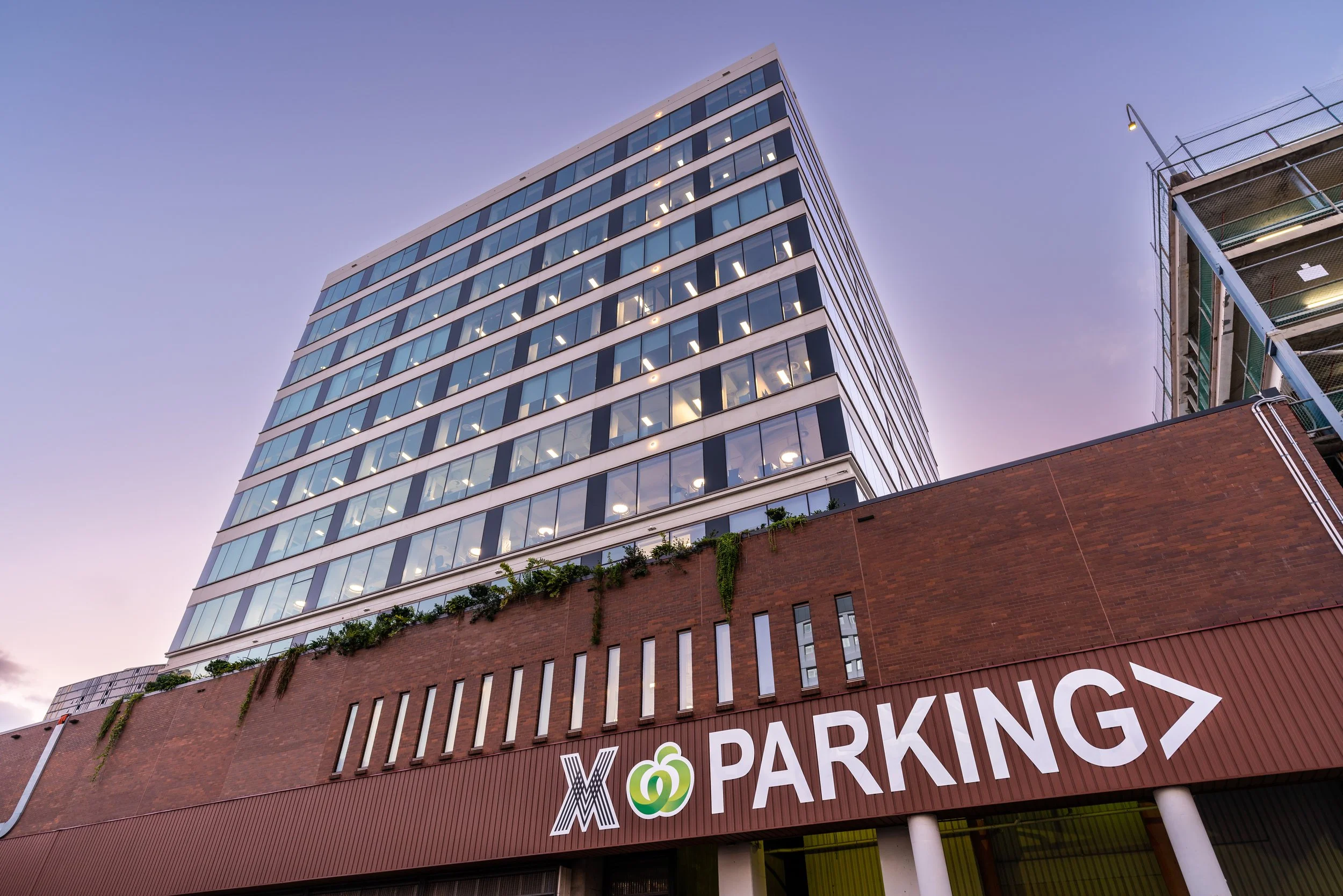 A modern multi-story office building with glass windows, viewed from below with a parking garage below it, and a large sign indicating parking and nearby parking options.
