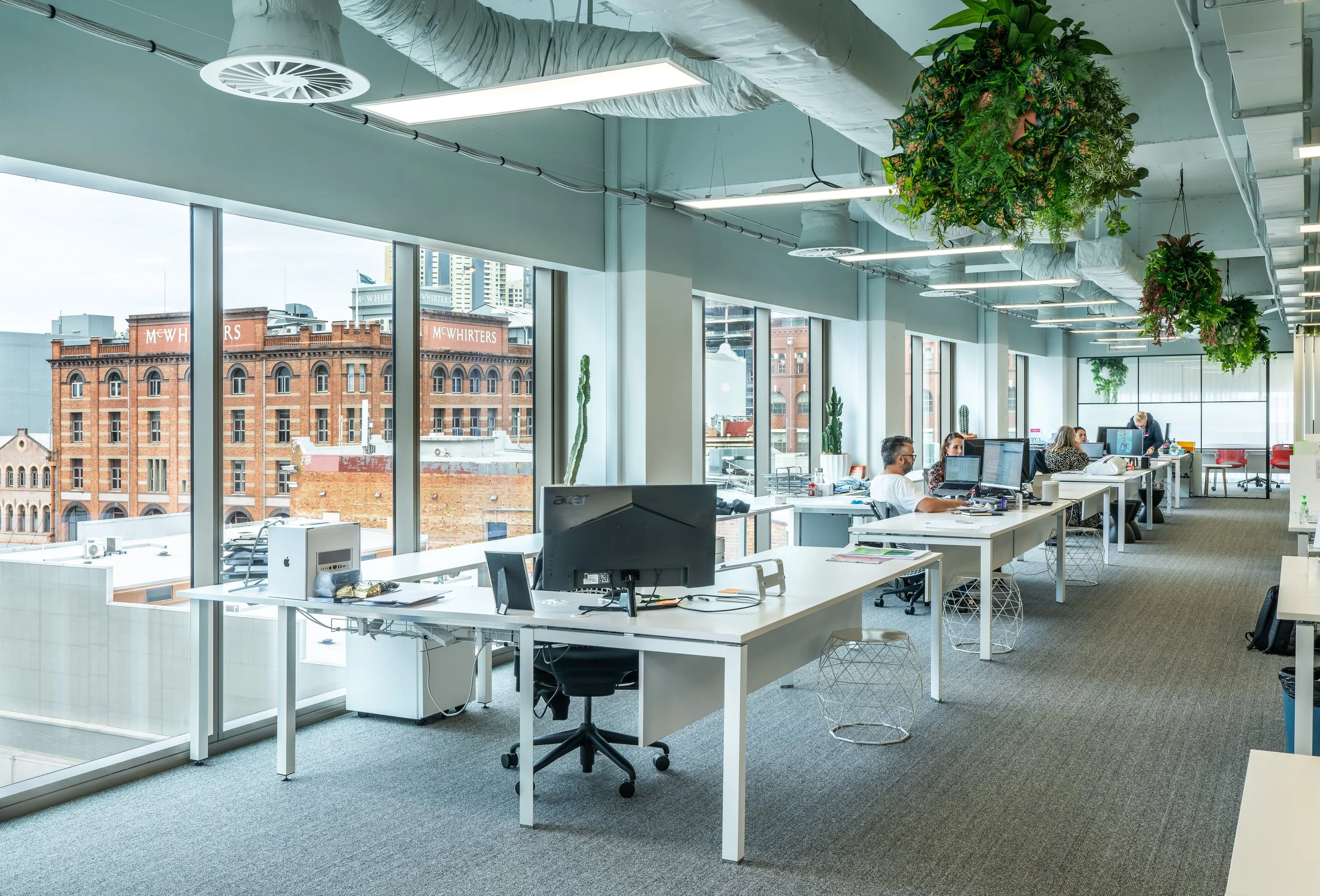Modern office space with large windows, white desks with computers, and hanging green plants, with a cityscape view outside.