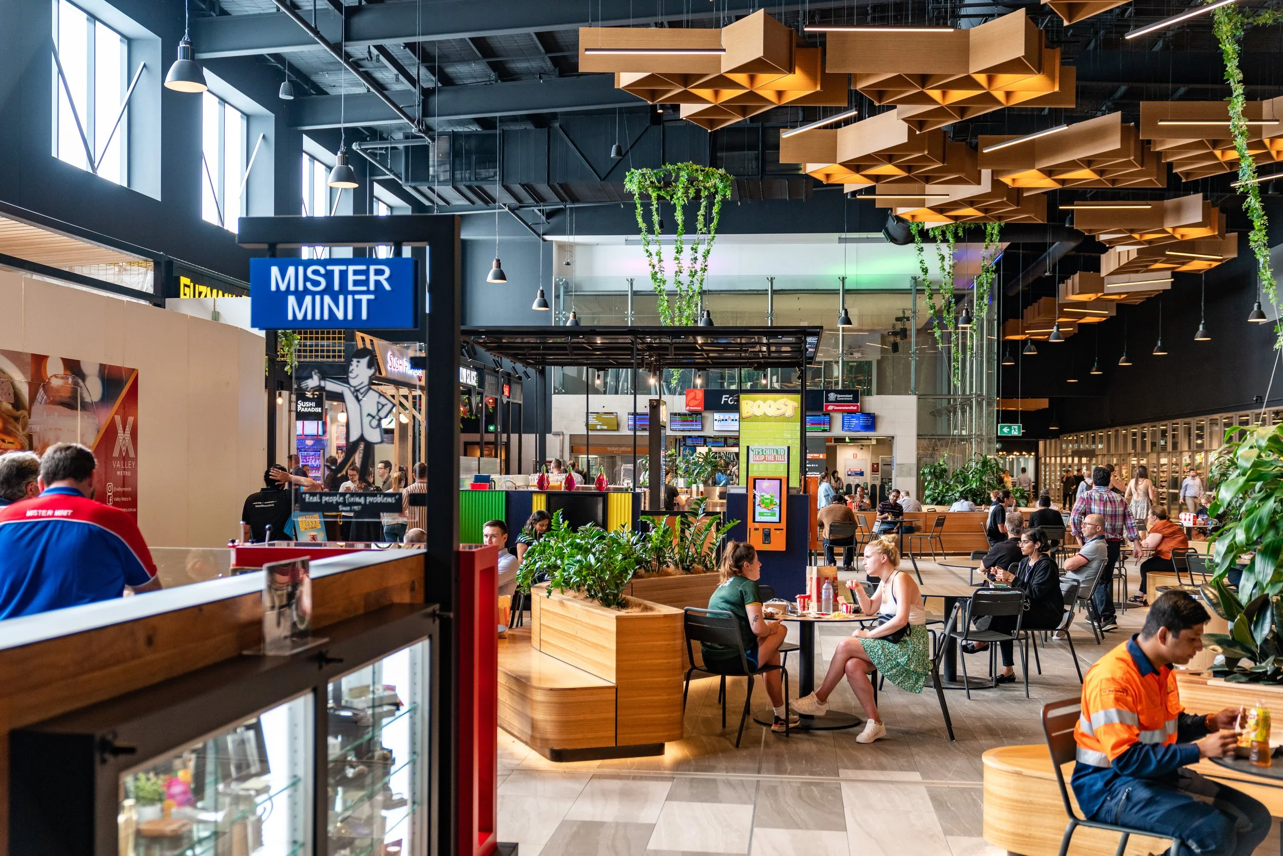 A busy indoor food court with people sitting at tables and walking around. There are various food stalls, greenery, and modern lighting fixtures.