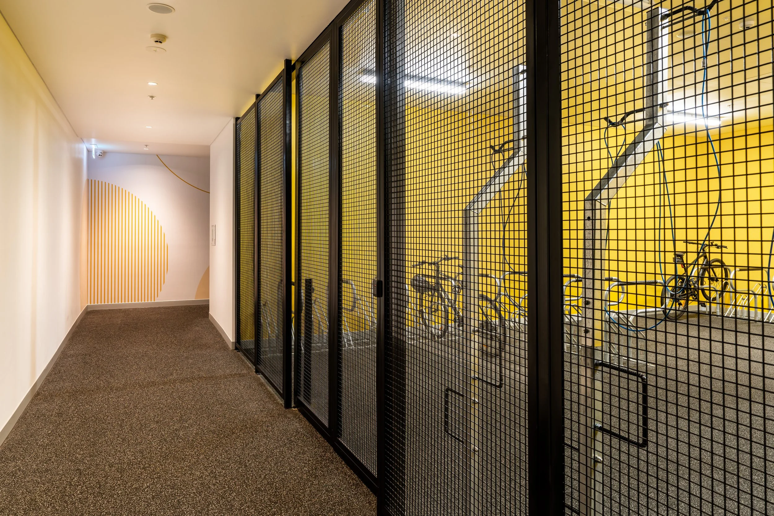 A corridor with a white wall and a yellow geometric mural on the left, and a glass enclosure with a yellow background containing several bicycles inside, separated by a black metal grid fence.