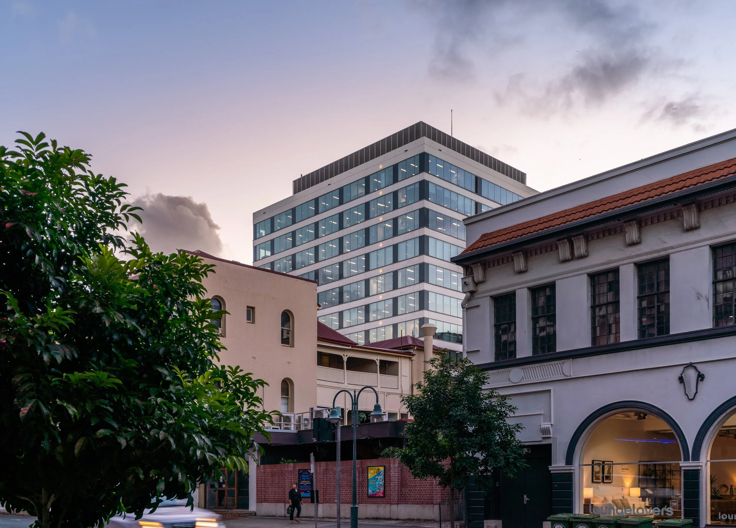 Street scene featuring a modern glass office building in the background, older white buildings in the foreground, trees, street lamps, and a person walking, during dusk with a cloudy sky.