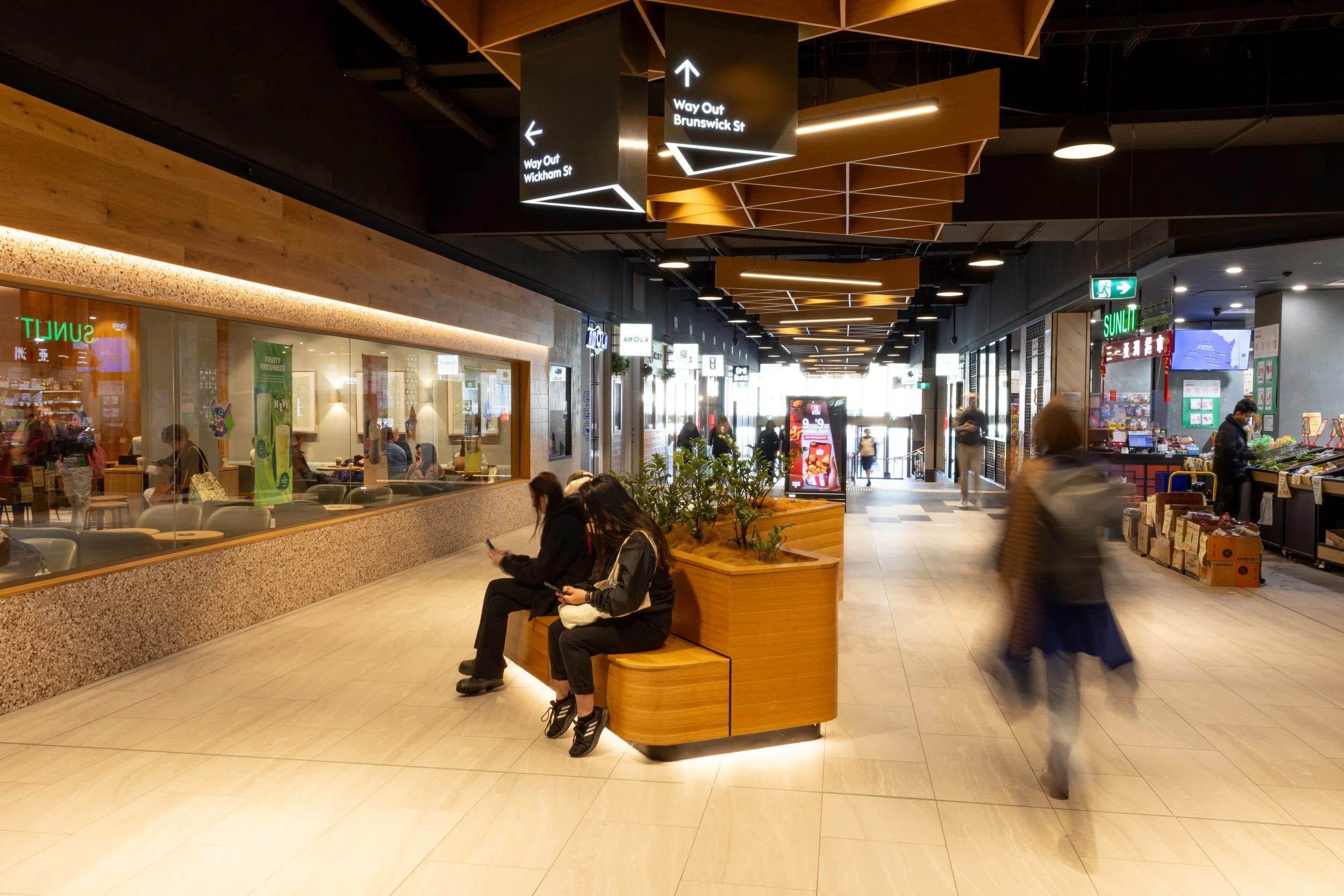 People sitting on a wooden bench with greenery in an indoor shopping mall, some using their phones, with signs for exits and shops visible overhead.