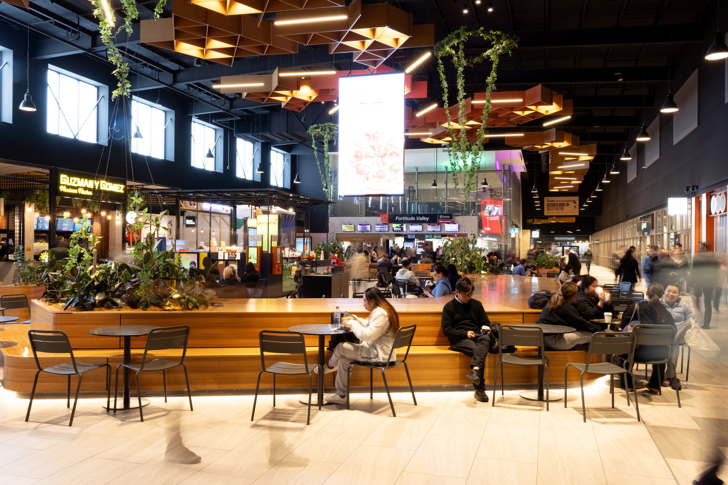 Inside a busy airport terminal with people sitting at tables and walking around. There are plants and modern ceiling decorations, signage for various shops and gates, including Guzmán y Gómez and Fortitude Valley.