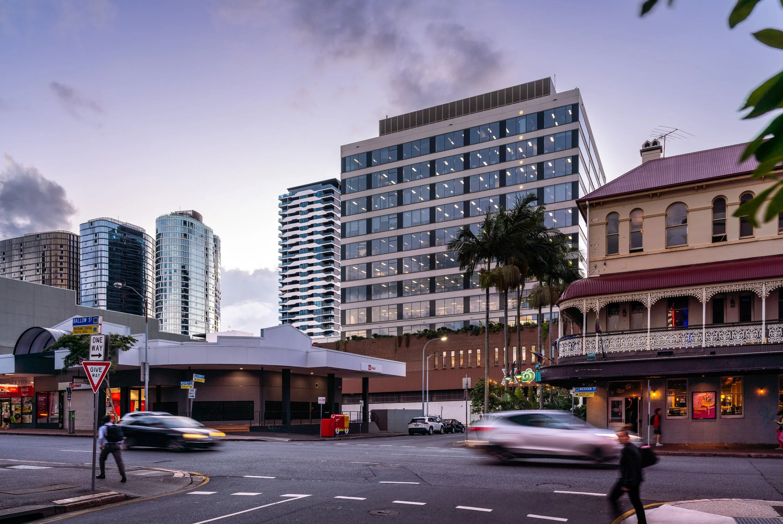 City street scene at dusk with modern high-rise buildings in the background, an older building with ornate balconies on the right, palm trees, and cars and pedestrians in motion.