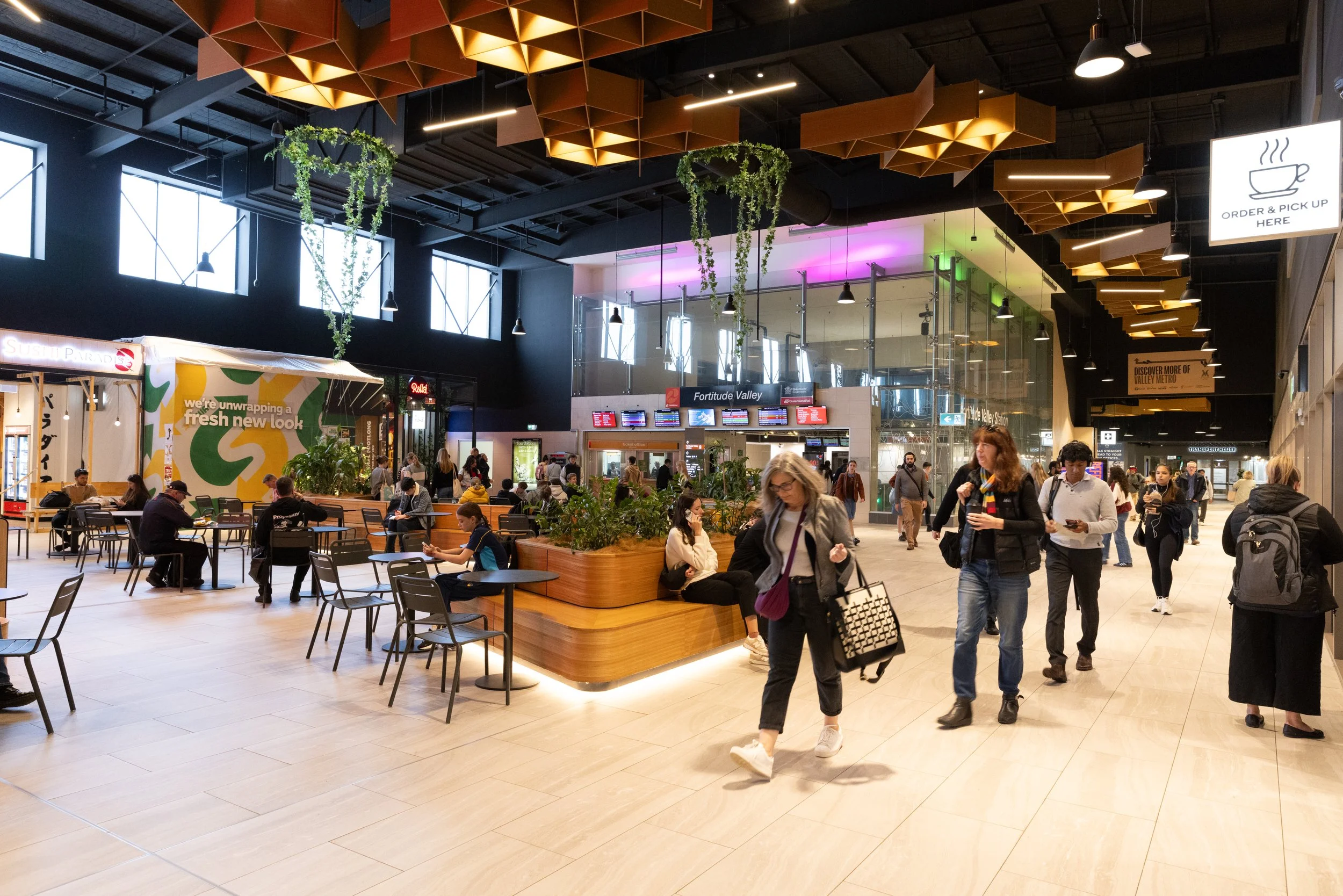 Interior of a modern shopping mall or transportation hub with people walking and sitting, a central waiting area with plants, an elevator, and signage including an order pickup sign.