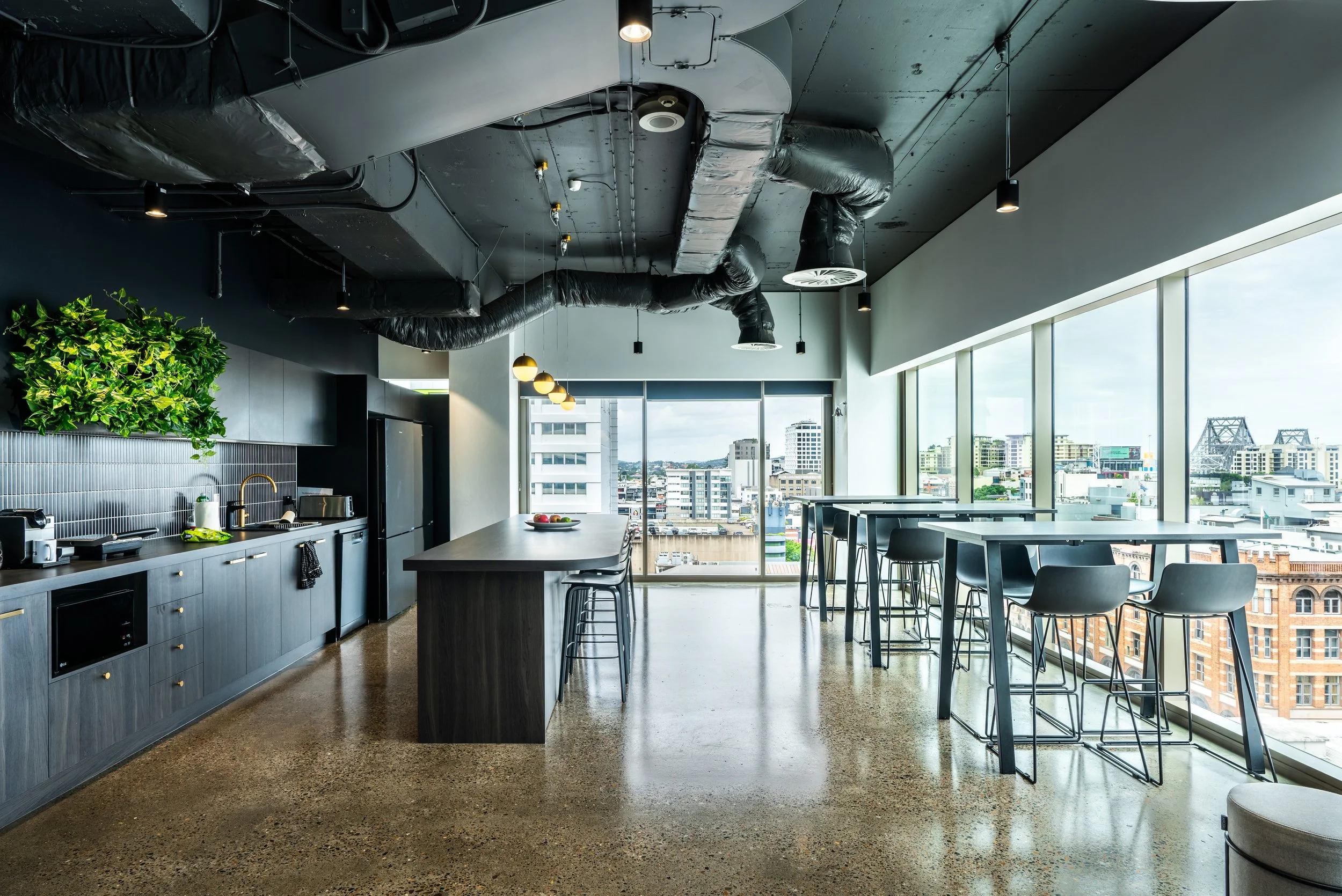 Modern high-rise apartment kitchen and dining area with large windows overlooking cityscape, featuring black cabinetry, kitchen appliances, and a high table with bar stools.