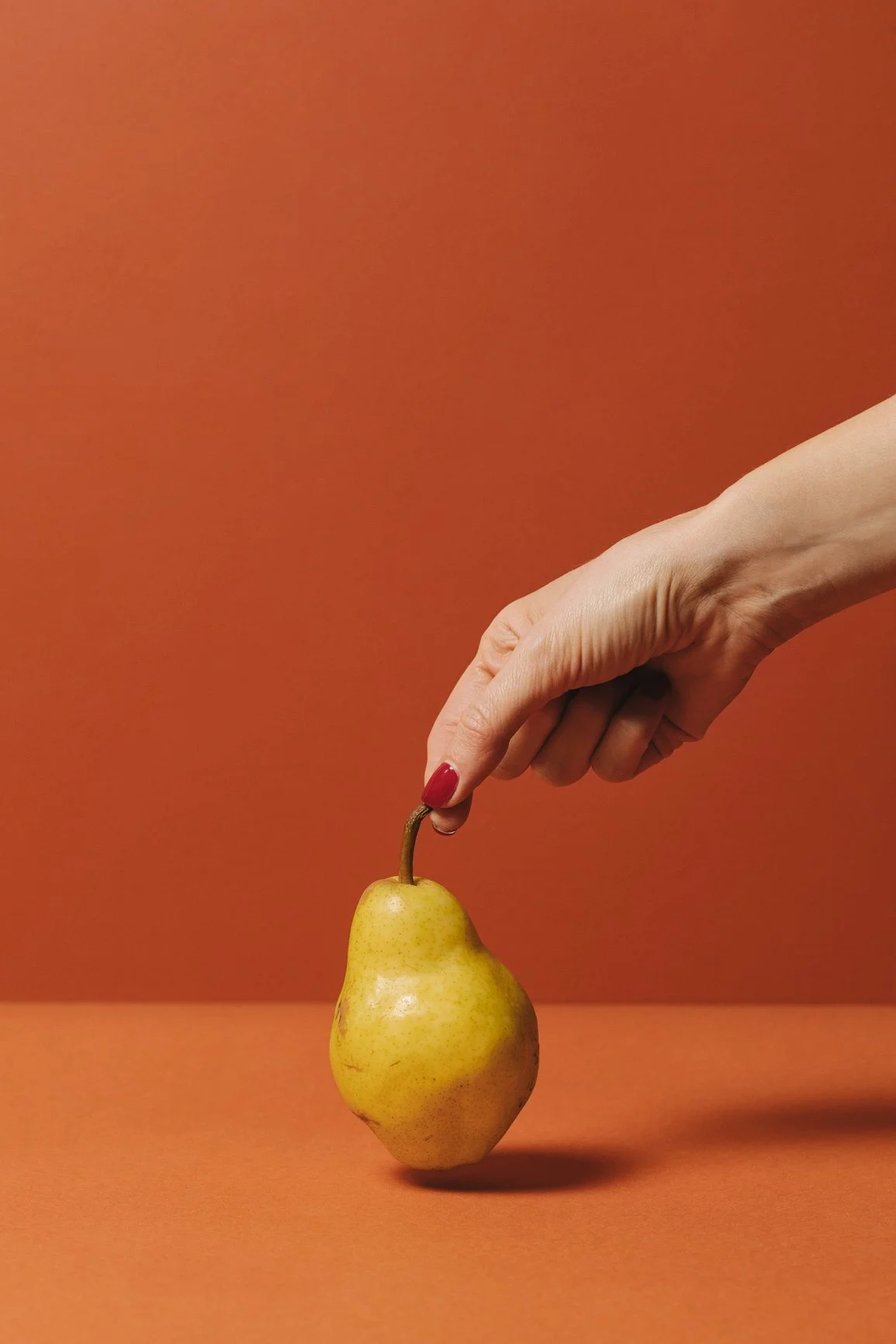 A hand with red painted nails holding the stem of a pear against a copper-colored background.