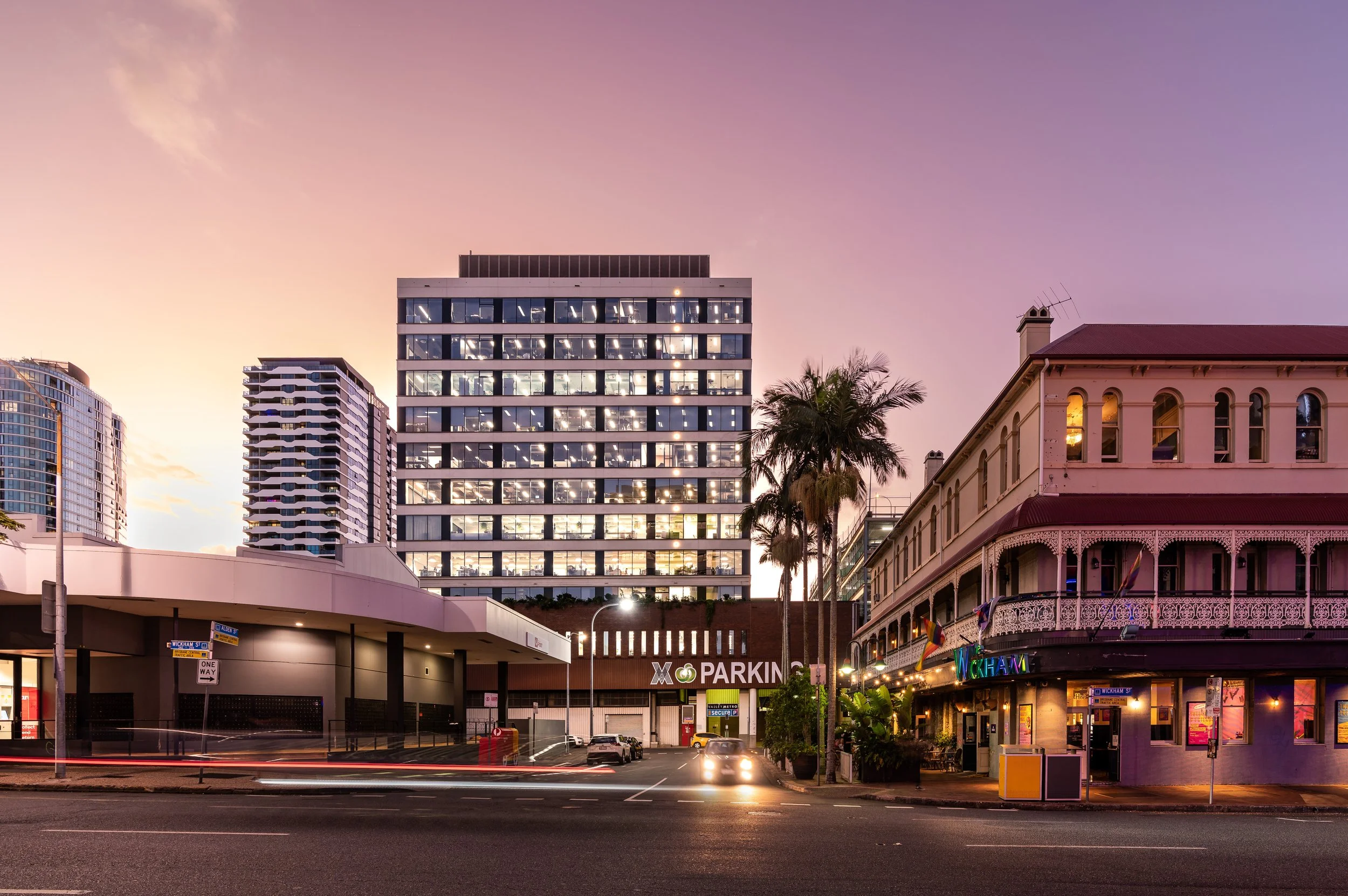City street scene at dusk featuring a modern glass building, a historic pink and cream building with outdoor seating, palm trees, car lights, and a parking garage entrance.