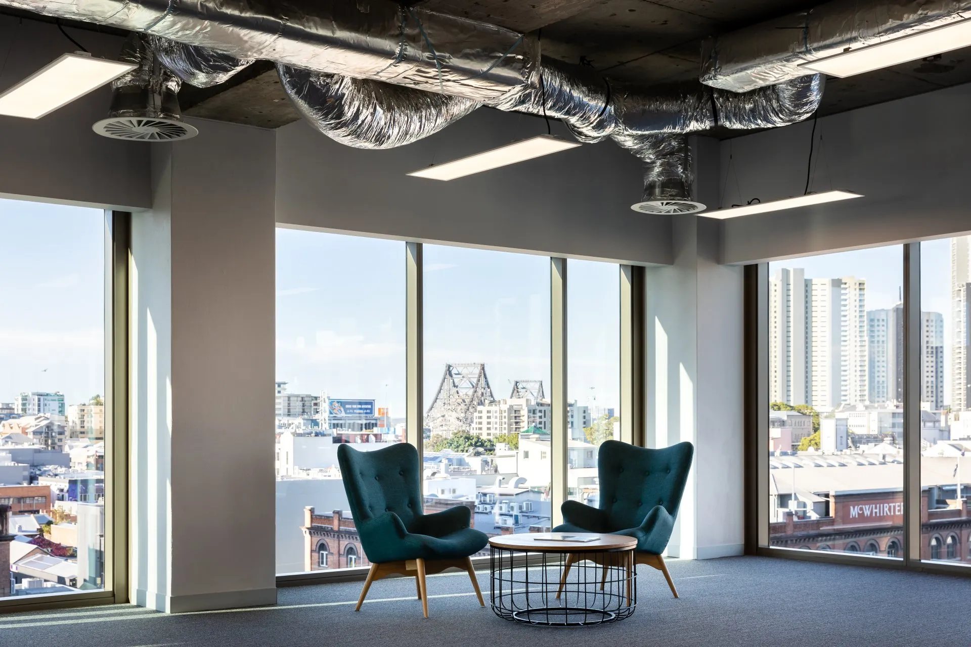 Office space with large windows, two teal armchairs, a round white table with books, and an unfinished ceiling with exposed ductwork.