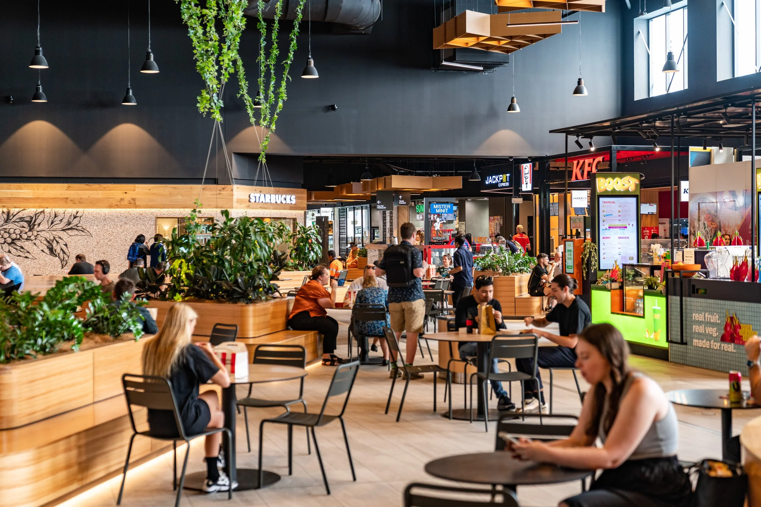 Interior of a modern shopping mall or food court with people sitting at tables, eating, using phones, and walking around near food stalls including Starbucks, KFC, and Boost. The area features wooden accents, greenery, and black ceiling decor with ha