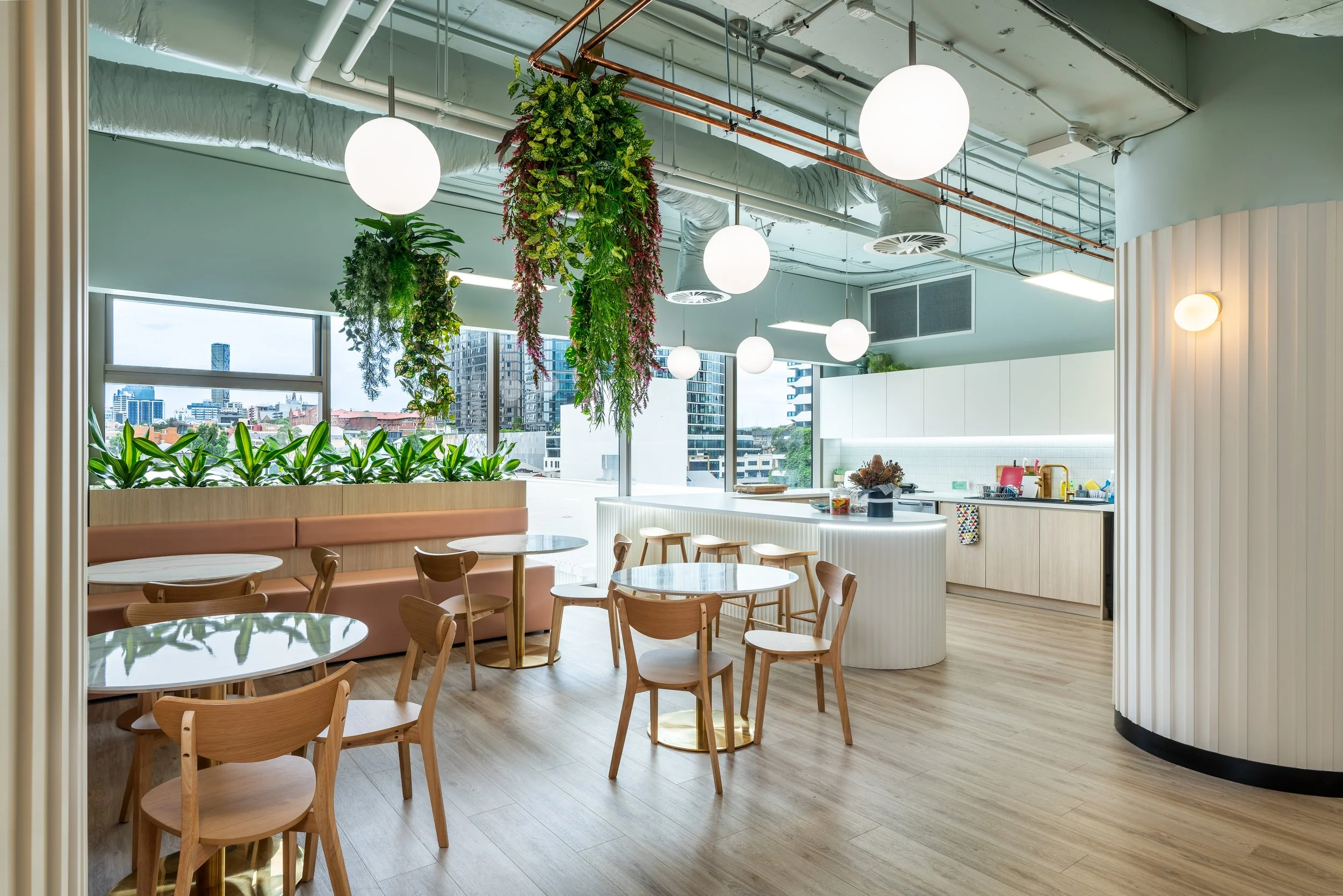 Bright, modern dining area with round glass tables, wooden chairs, a peach-colored cushioned bench, hanging greenery, and round white pendant lights. Large windows reveal a cityscape background.