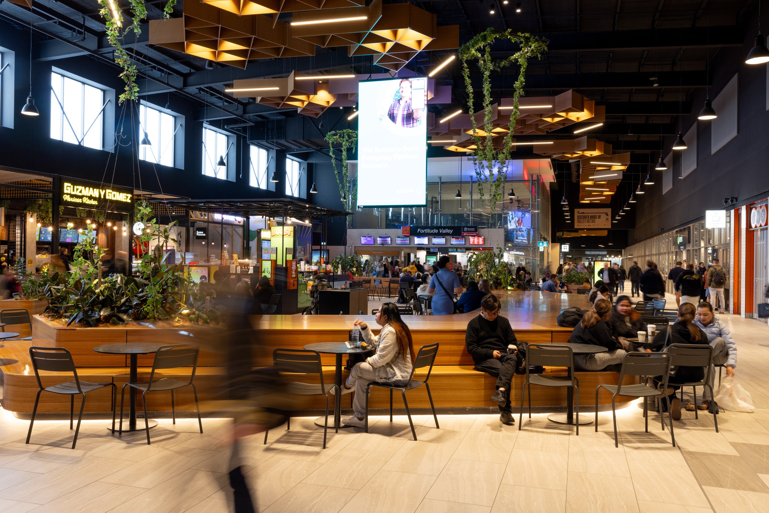 People sitting and walking in a modern indoor shopping mall with high ceilings, large windows, and various shops and food stalls.