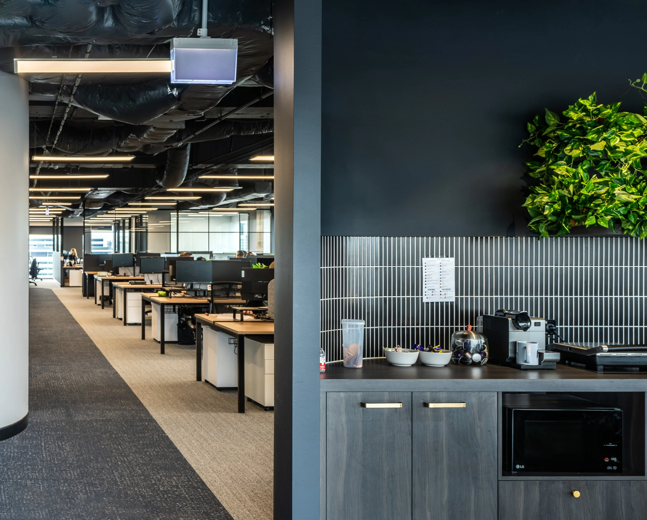 Office break area with coffee maker, bowls of snacks, and a plant on the counter, with open office desks visible in the background.
