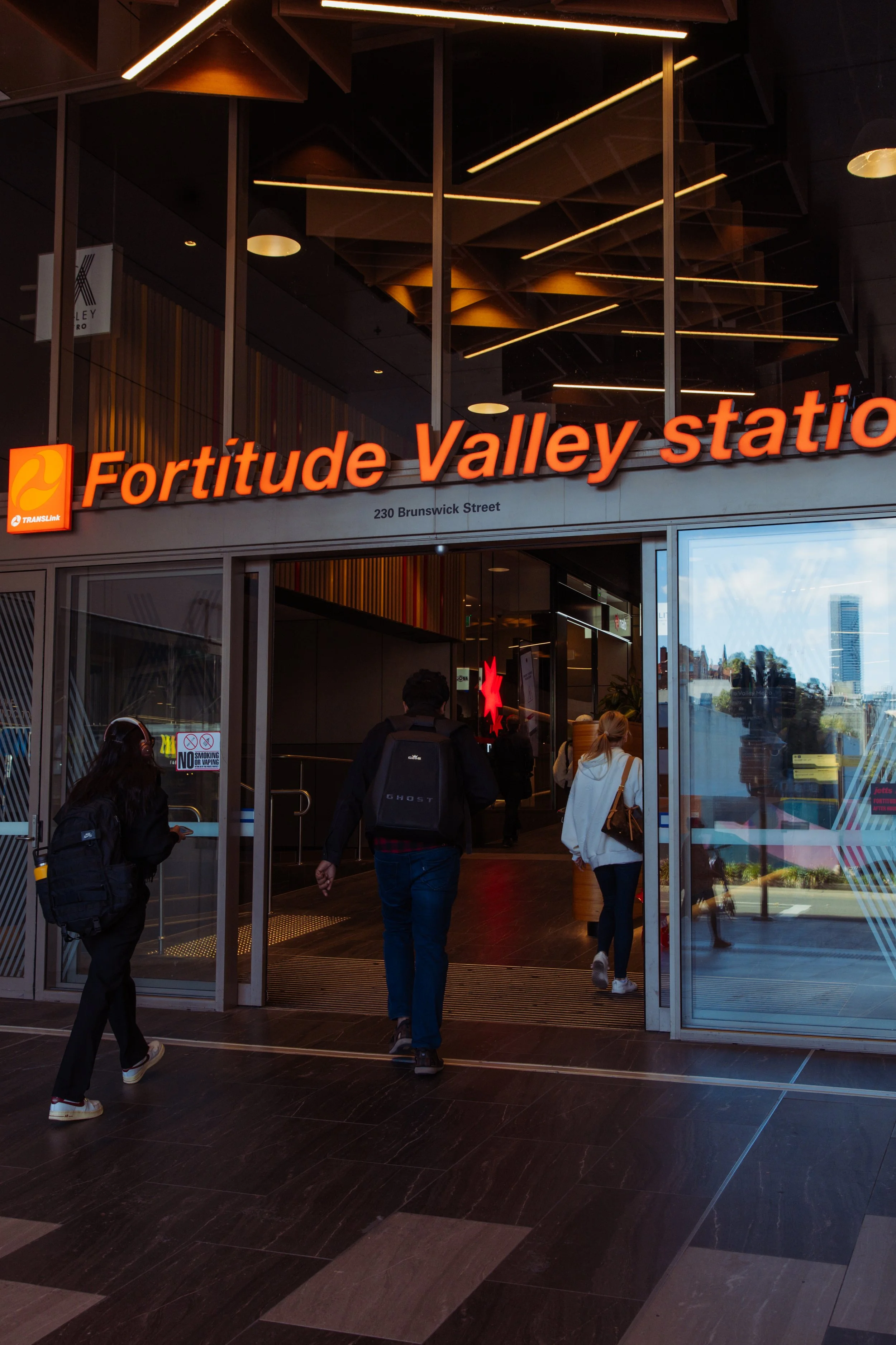 People entering the Fortitude Valley station with bright orange signage, modern interior, and reflective glass exterior.