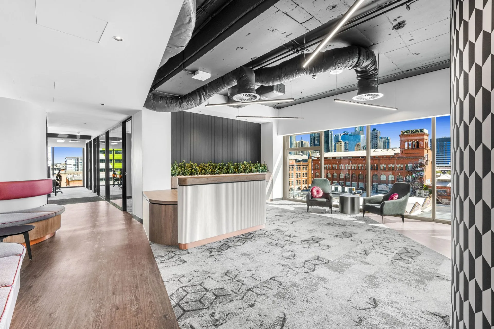 Modern office lounge area with large windows, city view, gray chairs with pink cushions, indoor plants, patterned rug, and exposed ceiling pipes.