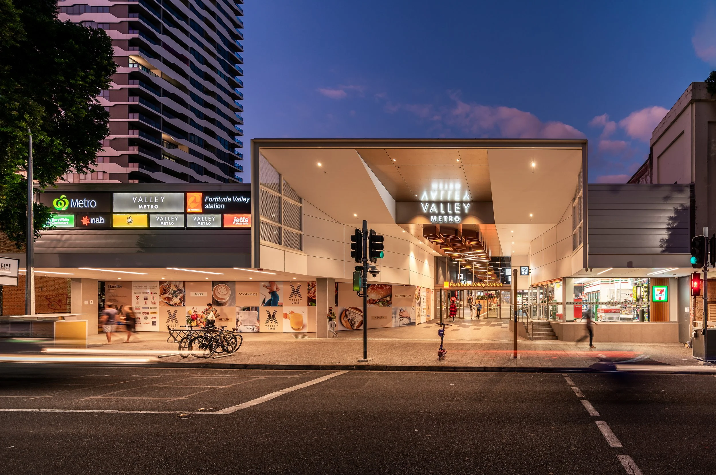 The Valley Metro station entrance at dusk, featuring modern architecture with illuminated signage and a clear evening sky. There are pedestrians, bicycles, and a scooter nearby, with shops and billboards visible around the station.