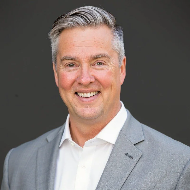 A smiling middle-aged man with gray hair, wearing a light gray suit and white shirt, standing against a black background.