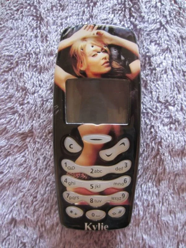 A black feature phone with an image of a woman with blonde hair on the screen. The phone has white number keys and the name 'Kylie' at the bottom.